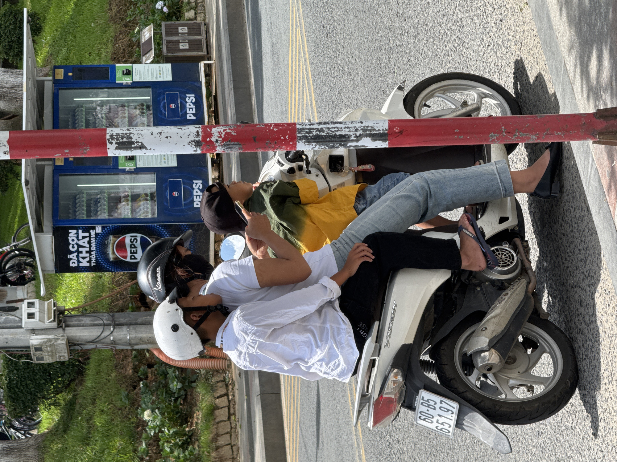 A group of people are riding a scooter in Da Lat, Vietnam. The scooter has a white body with silver accents. The license plate reads "60-B5 651.97". Three people are on the scooter. The person in the front is wearing a white helmet and a white long-sleeved shirt. They are holding a smartphone. The person in the middle is wearing a black helmet and a white t-shirt. They appear to be looking at the smartphone. The person in the back is wearing a black cap and a yellow and green jacket. They are looking forward. In the background, there is a Pepsi vending machine and a red and white striped pole. The ground is asphalt. There are trees and grass on the left side of the image. The sky is bright and sunny.