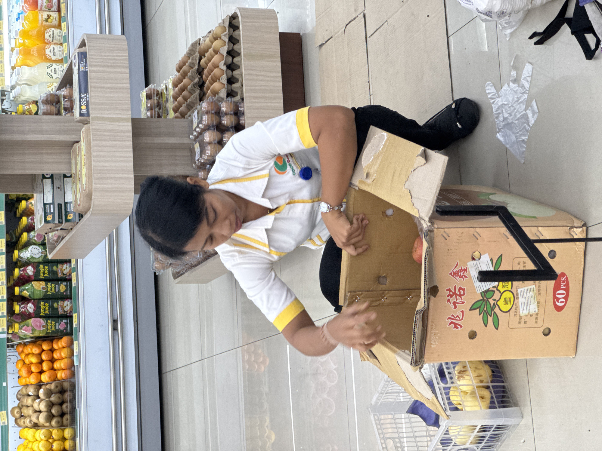 A woman in a white shirt with yellow trim and black pants is kneeling on the floor of a supermarket in Kuta Selatan, Indonesia. She is sorting through a cardboard box of fruit. The box has Chinese writing on it and a "60 PCS" label. To her left is a white plastic basket containing more fruit. In the background, there are shelves stocked with various products, including juices, kiwis, oranges, and cartons of eggs. The floor is tiled.