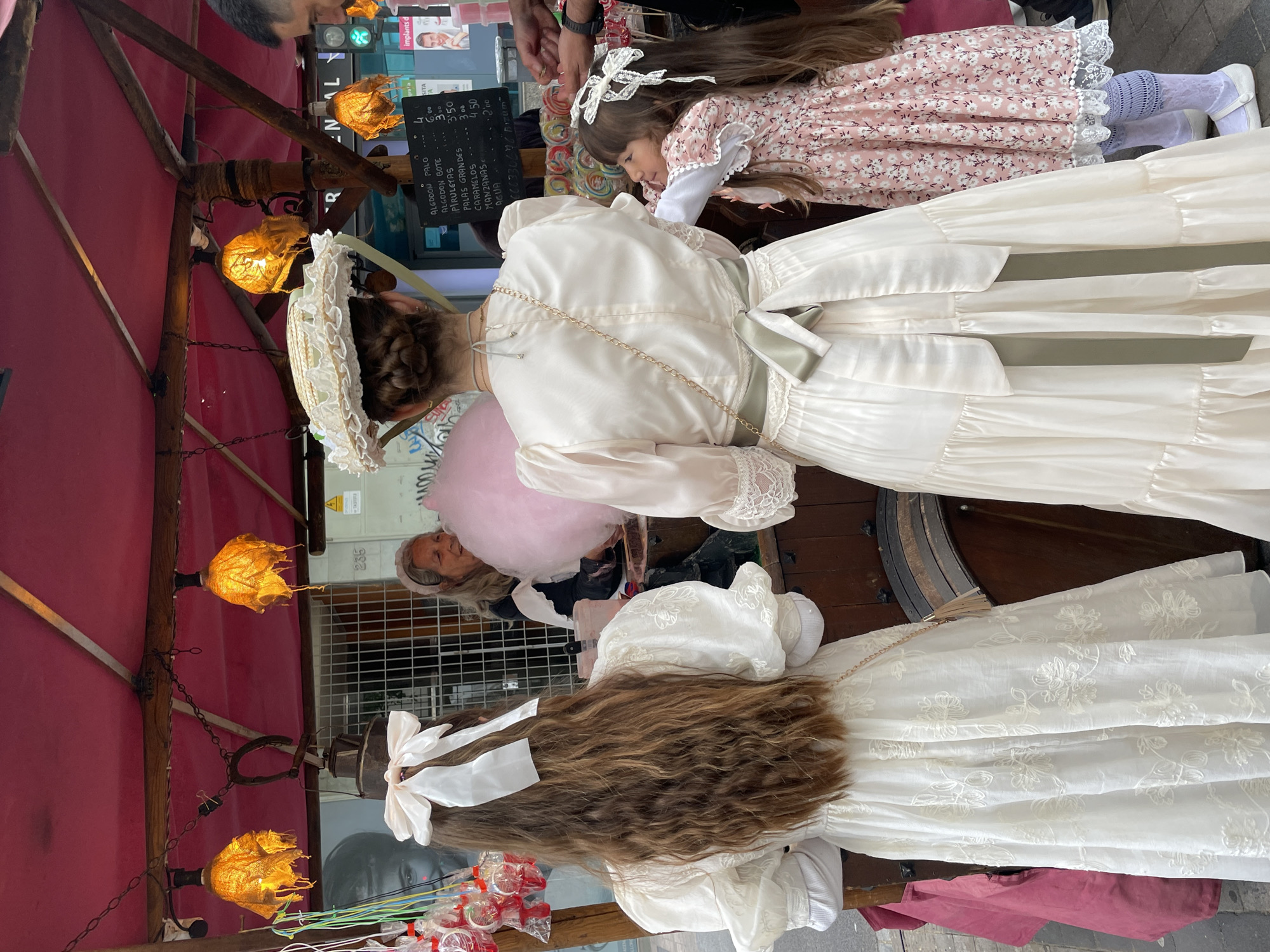This image captures a scene at an outdoor market or fair, possibly in Terrassa, Spain. Three girls are wearing old-fashioned dresses with lace and ribbons, which are indicative of a historical reenactment or special event. One girl wears a white bonnet. An elderly woman with a gray headband holds a large pink cotton candy, while a vendor's stall is visible with lanterns hanging above and a menu board listing prices for items like "Algodon Palo" (cotton candy on a stick), "Piruletas" (lollipops), and "Manzanas" (apples), with the option to pay via "Bizum." Lollipops are also visible on display. The stall has a red awning, and parts of street signs and building facades are visible in the background.