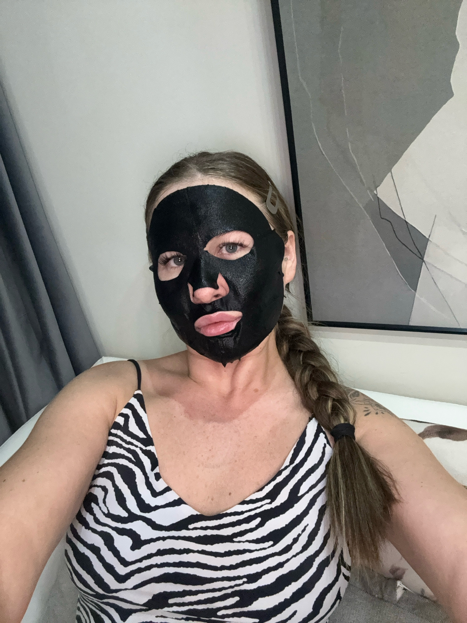In a close-up, selfie-style shot, a woman with long, braided blonde hair is shown from the chest up, wearing a black sheet mask and a black and white zebra-striped camisole. Her eyes are visible through cutouts in the mask, and her lips are slightly parted. The background is neutral, with a gray curtain on the left and a framed abstract artwork on the right. The overall setting appears to be indoors.