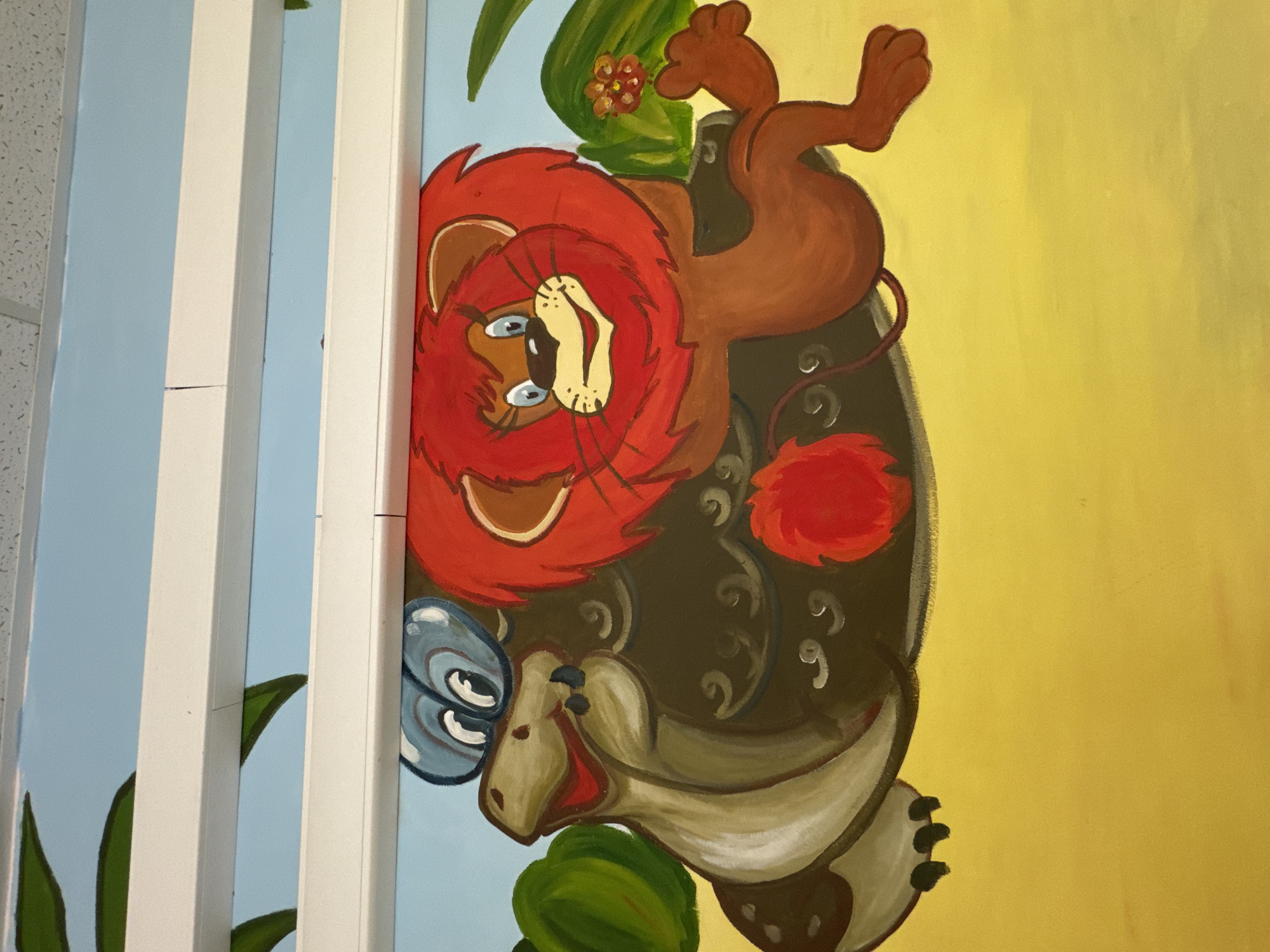 The image is a close-up, horizontal, eye-level photograph of a mural painted on a wall. The mural depicts cartoon animals in a savanna-like environment. On the left, a tan-colored creature resembling a turtle or dinosaur with large blue glasses is shown. It has a long neck, an open mouth showing red and white, and dark spots on its body. Beside it, a brown creature with swirling patterns on its back, likely the turtle's shell, is visible. To the right, a cartoon lion with a bright red mane and a yellow face is featured. The lion has blue eyes, a smiling expression, and is holding a small orange flower in its right paw. Its body is brown, and it is depicted in a seated or lying position. Red, fluffy shapes, possibly flowers or pom-poms, are also present on the mural. The background consists of a light blue sky and a yellow ground. Green foliage is visible in the upper left and right portions of the mural. Two white, horizontal bars are positioned across the top of the image, partially obscuring the mural. The ceiling tiles are visible above these bars. This mural is located in Vidnoye, Russia, likely on a wall in a public space or children's area.