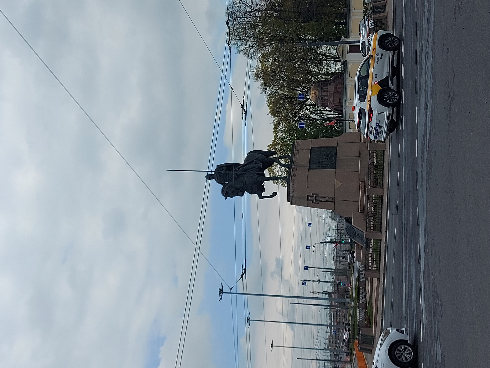 The image depicts the equestrian statue of Nicholas I in St. Petersburg, Russia. The statue is mounted on a granite pedestal and depicts Nicholas I on horseback. A street runs alongside the statue with taxi cabs parked on the side of the road. Overhead wires are strung along the street. Trees are visible behind the statue. The sky is cloudy.