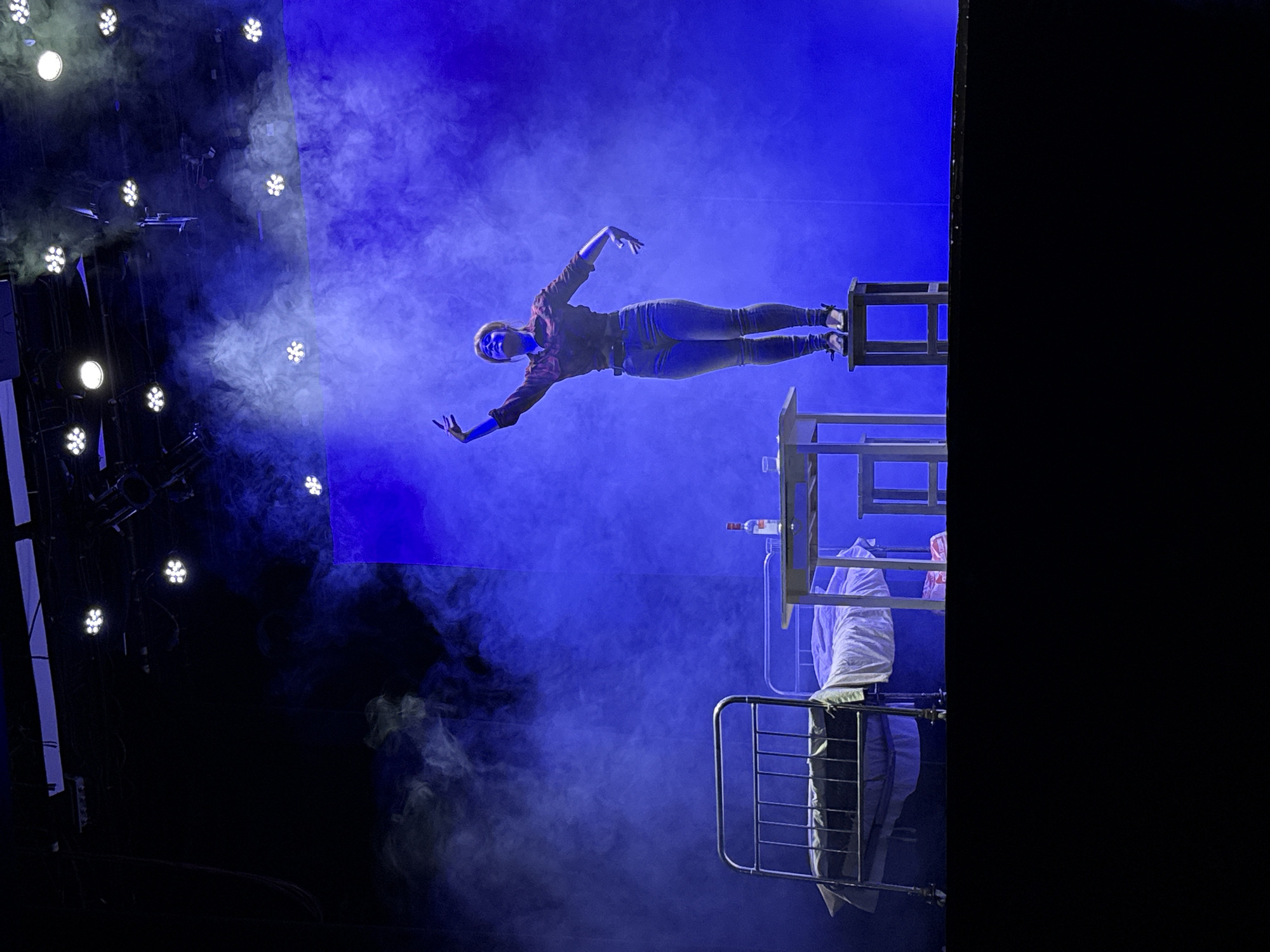 The image shows a stage performance, likely a dance or theatrical production, in a dark setting illuminated by blue lighting and haze. A person is positioned horizontally, suspended on a stack of wooden chairs. The person is wearing dark jeans and a long-sleeved shirt. To the right of the person is a metal bed frame, draped with white cloth. Above the bed frame, resting on the chairs, is a bottle. The background features theatrical lighting and fog effects. Given the lighting and staging, the location is likely an indoor performance space in Chaikovsky, Russia.