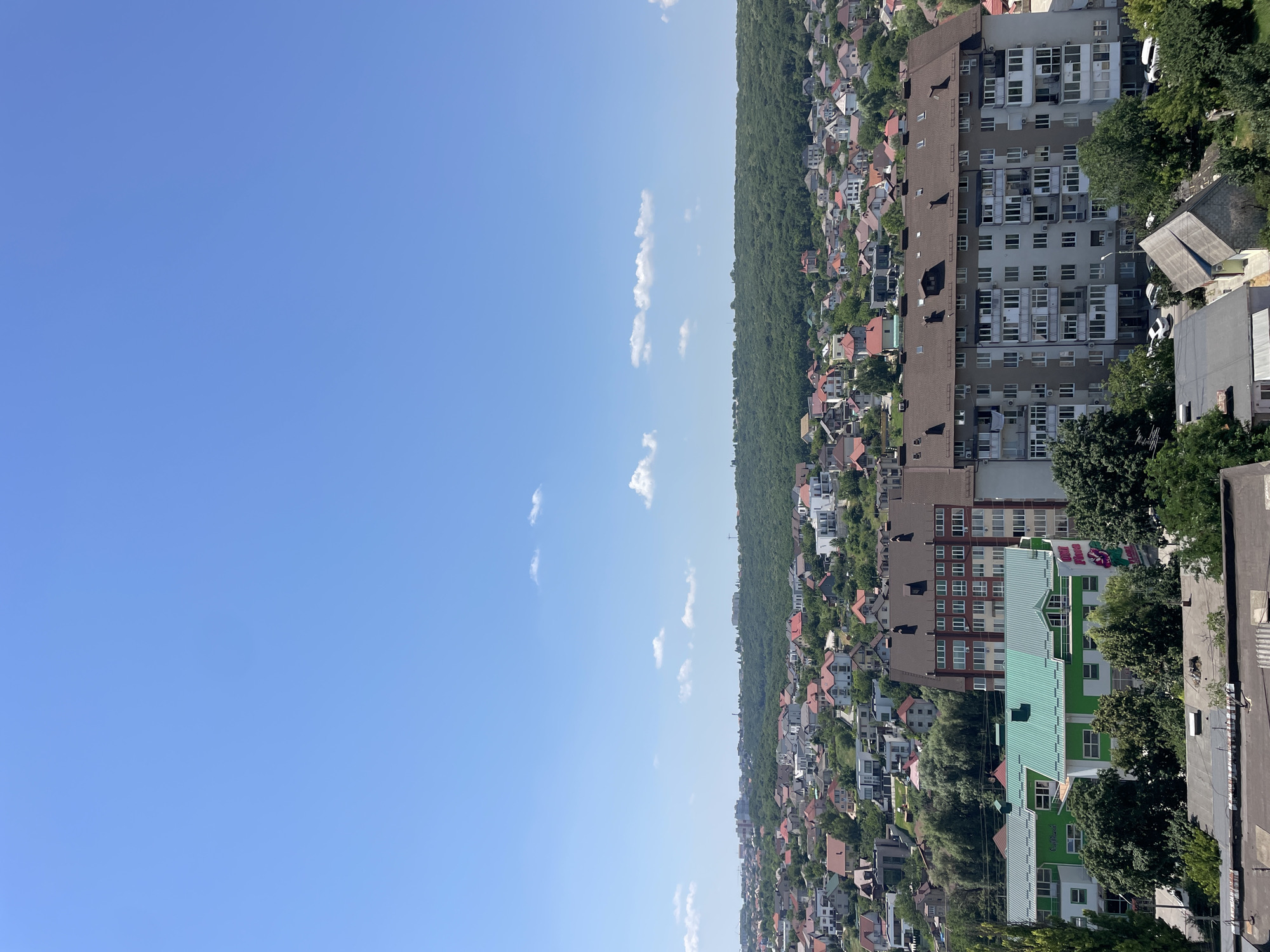 This is a medium-angle, high-altitude, eye-level shot of a sprawling residential area under a clear blue sky with scattered white clouds. In the foreground, several flat-roofed structures are visible, with one in the lower left corner appearing to be made of corrugated metal. A light-colored apartment building with multiple stories and numerous balconies dominates the mid-ground. To the left of this apartment building is a lower, bright green building with a pitched roof. Behind these structures, a dense network of houses with red, orange, and gray roofs extends into the distance, interspersed with trees. A thick band of green forest covers the horizon, with a few taller buildings peeking through from the far left. The scene suggests a suburban or urban fringe setting on a sunny day.