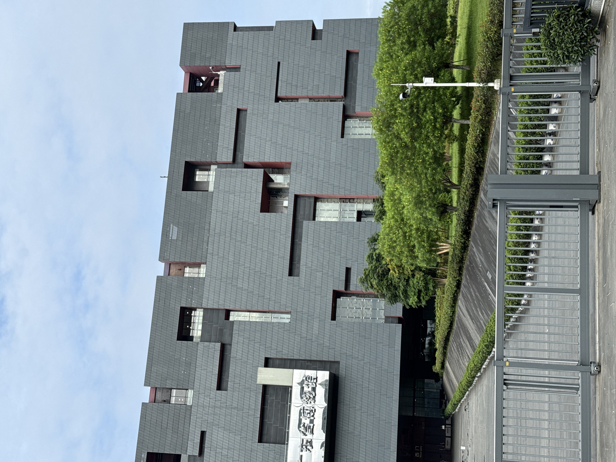 The image shows a tall, gray building with a modern, asymmetrical design in the Tianhe District, China. The building appears to be made of textured gray panels, with irregularly placed window openings and narrow red accent strips. There is Chinese lettering on a sign at the lower part of the building. To the right of the building is a tree-lined area with lush green foliage. In the foreground, there is a gray metal gate. The sky is partially visible, with light blue areas and a few clouds. A security camera is mounted on a pole near the gate. The image is taken from a perspective that makes the building appear tilted.