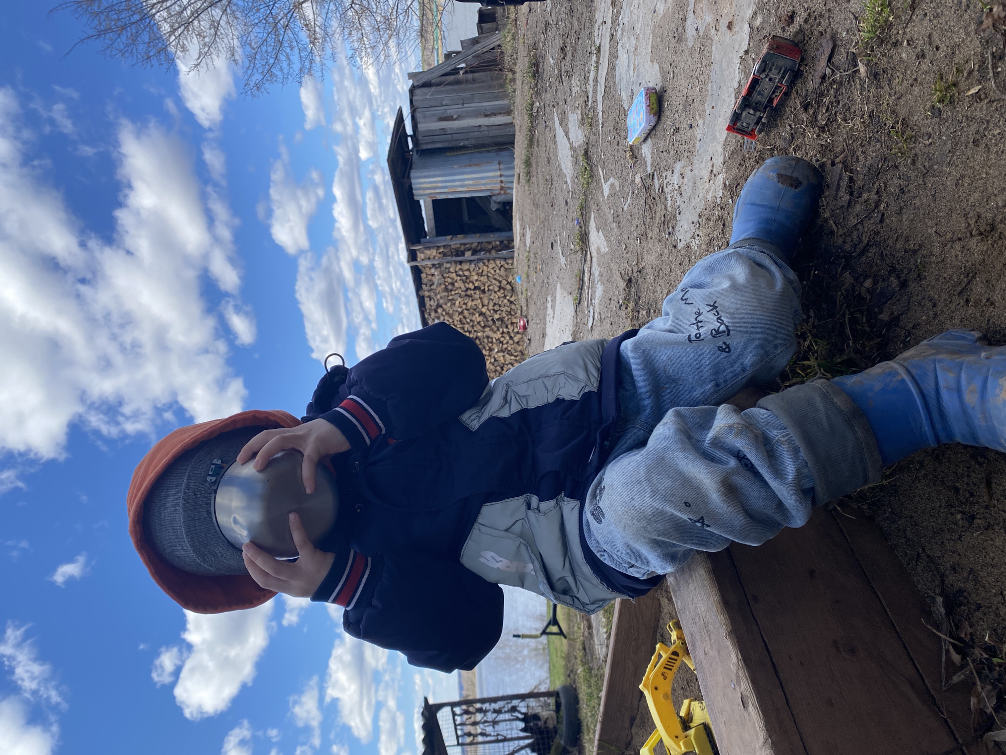 A child wearing a rust-colored hood, a dark blue jacket with light gray panels, light blue jeans, and blue rubber boots is lying on the ground outdoors. The child is holding a shiny metal bowl over their face.
In the background, there's a wooden structure with stacks of firewood. There are toy construction vehicles and a toy car nearby. The background also shows a blue sky with white clouds and some greenery. In the distance, there is a body of water and a fence.
The location is potentially the city of Ольховка, Russia.