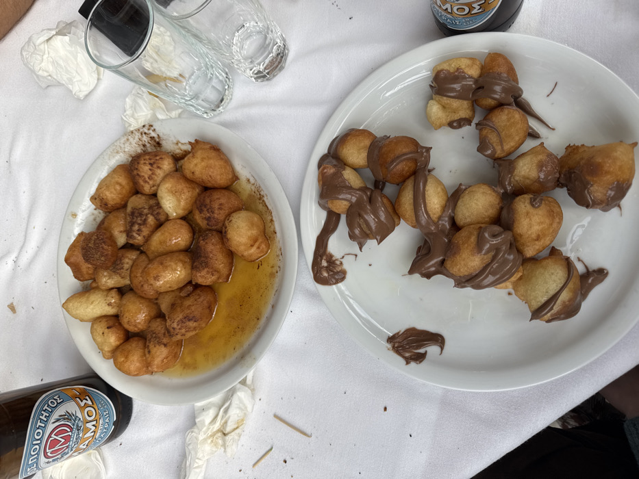 The image provides an overhead view of a dining table located in Nea Smirni, Greece. The table is covered with a white tablecloth. Two white ceramic plates of Loukoumades (Greek donuts) are centrally positioned. The upper plate contains approximately 25-30 golden-brown fried dough balls partially submerged in a golden-brown syrup, likely honey, with visible cinnamon dusting. A brown glass bottle, labeled 'ΜΠΕΛΟΠΟΙΟΤΗΣ ΣΑΜΟΣ' (Peloponnesian Samos), stands to the upper left of this plate. To the right of the upper plate are two clear glass tumblers of differing heights and several crumpled white paper napkins. The lower plate holds approximately 20-25 similar golden-brown fried dough balls extensively covered with a thick, dark brown, chocolate-hazelnut spread. A dark-colored beverage container with a partial blue label is visible to the right of the lower plate. Two small wooden toothpicks and minor food debris are scattered on the tablecloth, indicating recent consumption. No people are visible.