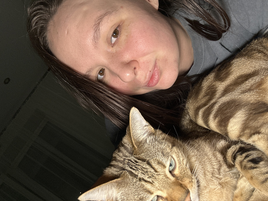 An indoor scene features a female individual and a domestic cat. The female, positioned on the right, has dark brown hair, light skin, and brown eyes, looking directly towards the camera with a slight pout. She is wearing a grey crew-neck garment. The cat, a brown tabby with striped fur and light green eyes, occupies the left side of the frame, looking towards the left with a somewhat disgruntled expression. The female's right arm or shoulder is visible underneath the cat. The environment is dimly lit, suggesting an interior space, possibly a residential setting. No specific location within the city of Chaikovsky, Russia, can be determined from the visual evidence.