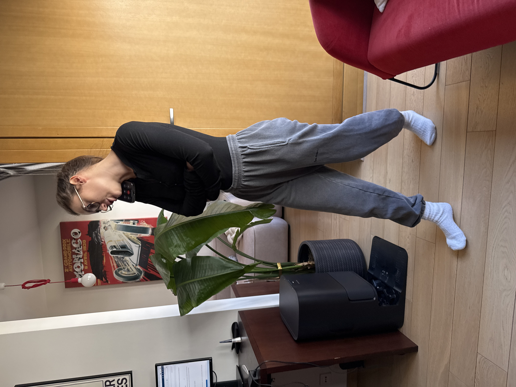 A woman is lying on her side on a wooden floor, talking on a cell phone. She is wearing glasses, a black long-sleeved shirt, gray sweatpants, and white socks. A red chair is visible in the background. To her left is a large potted plant next to a black printer on a wooden desk. Behind the plant is a poster depicting a race car with the words "MONACO" and "2e GRAND PRIX AUTOMOBILE MONACO 6 AVRIL 1930". There's also a framed picture on the wall. The location appears to be an indoor space, likely an apartment in Warsaw, Poland.