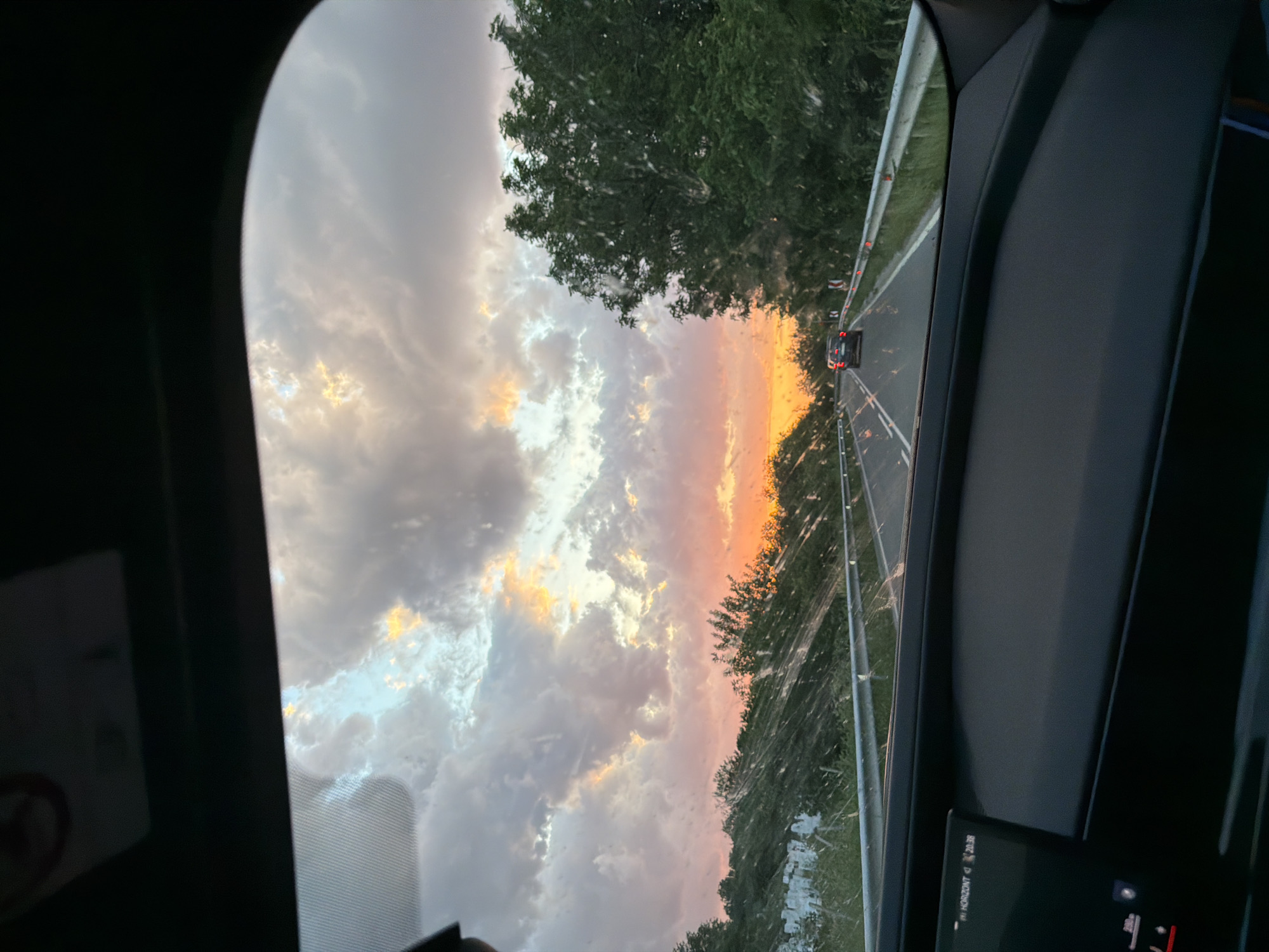 The image shows a view from inside a vehicle, likely a car, driving on a road. The road is bordered by trees and a guardrail. In the distance, a car is visible ahead on the road. The sky is filled with clouds, displaying a sunset with orange and pink hues. The location is possibly Novakovo, Bulgaria, based on the provided information, but it is impossible to confirm without additional context or landmarks.