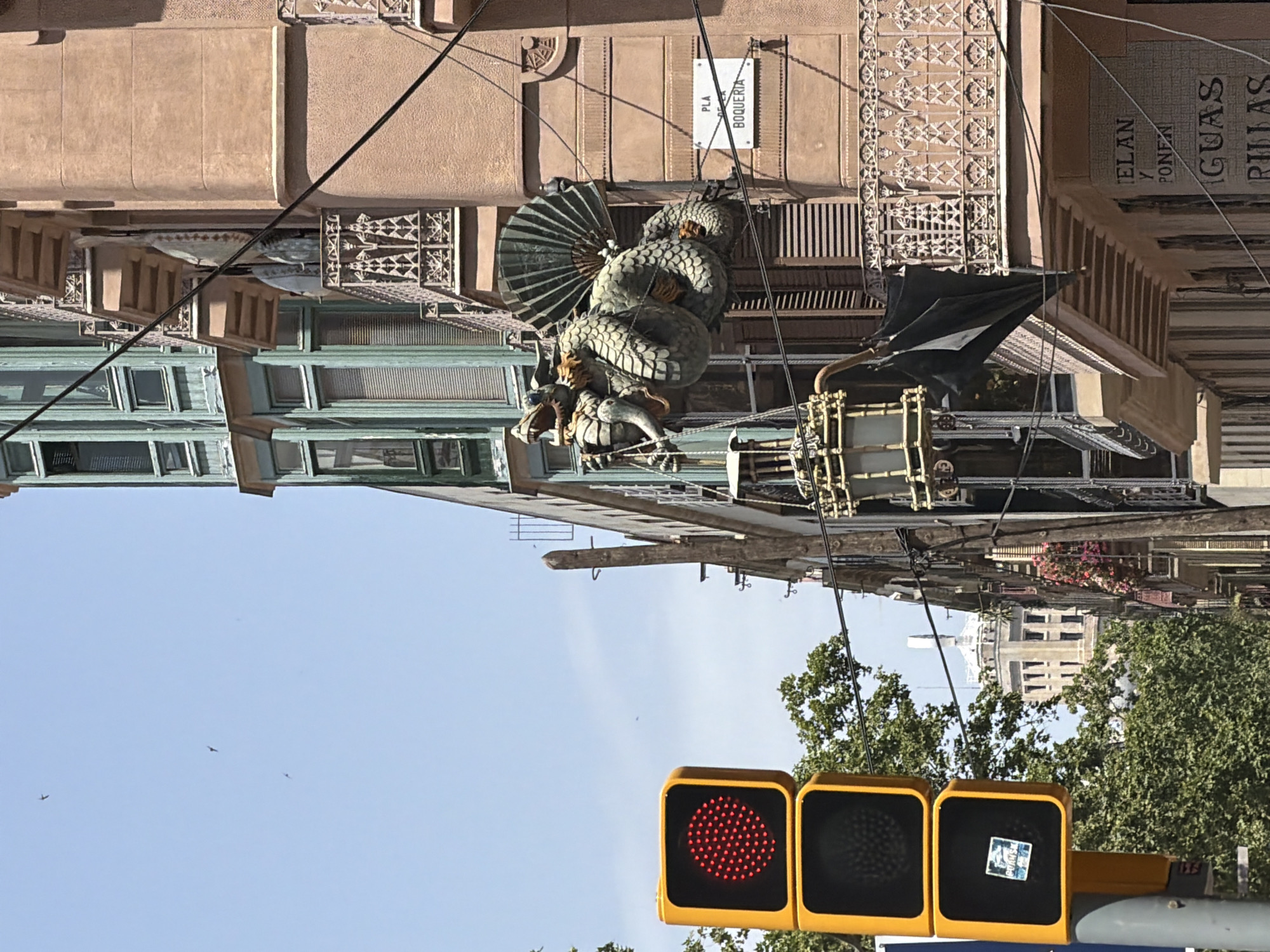 This is a photograph of a street in Barcelona, Spain, near Plaça de la Boqueria. In the foreground, a traffic light with a red signal is visible on the left. To the right of the traffic light, a building with a salmon-pink facade is prominent. Attached to the building's facade is a large, decorative sculpture of a green dragon, which appears to be coiled around a fan-like structure. A black umbrella is hanging from a balcony on the building below the dragon sculpture. Further down the street, other buildings and trees can be seen. The sky is clear and blue, with a few birds in flight. The street appears to be illuminated by daylight.