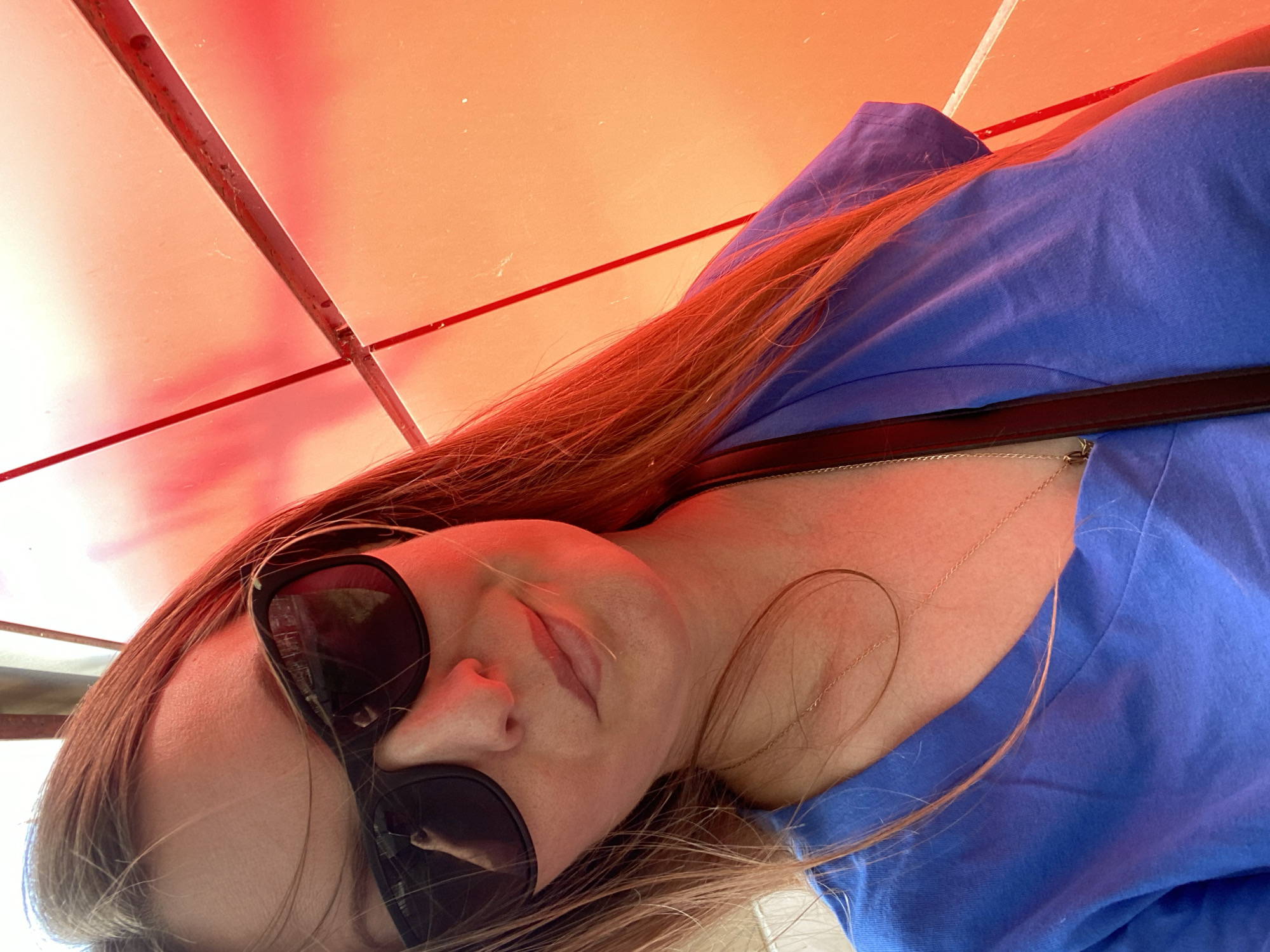 A woman is lying down, wearing sunglasses and a blue shirt. She has light skin and light brown hair. A black purse strap is visible across her chest. Above her is a red-orange structure, possibly a canopy or awning. The location is not discernible from the image.