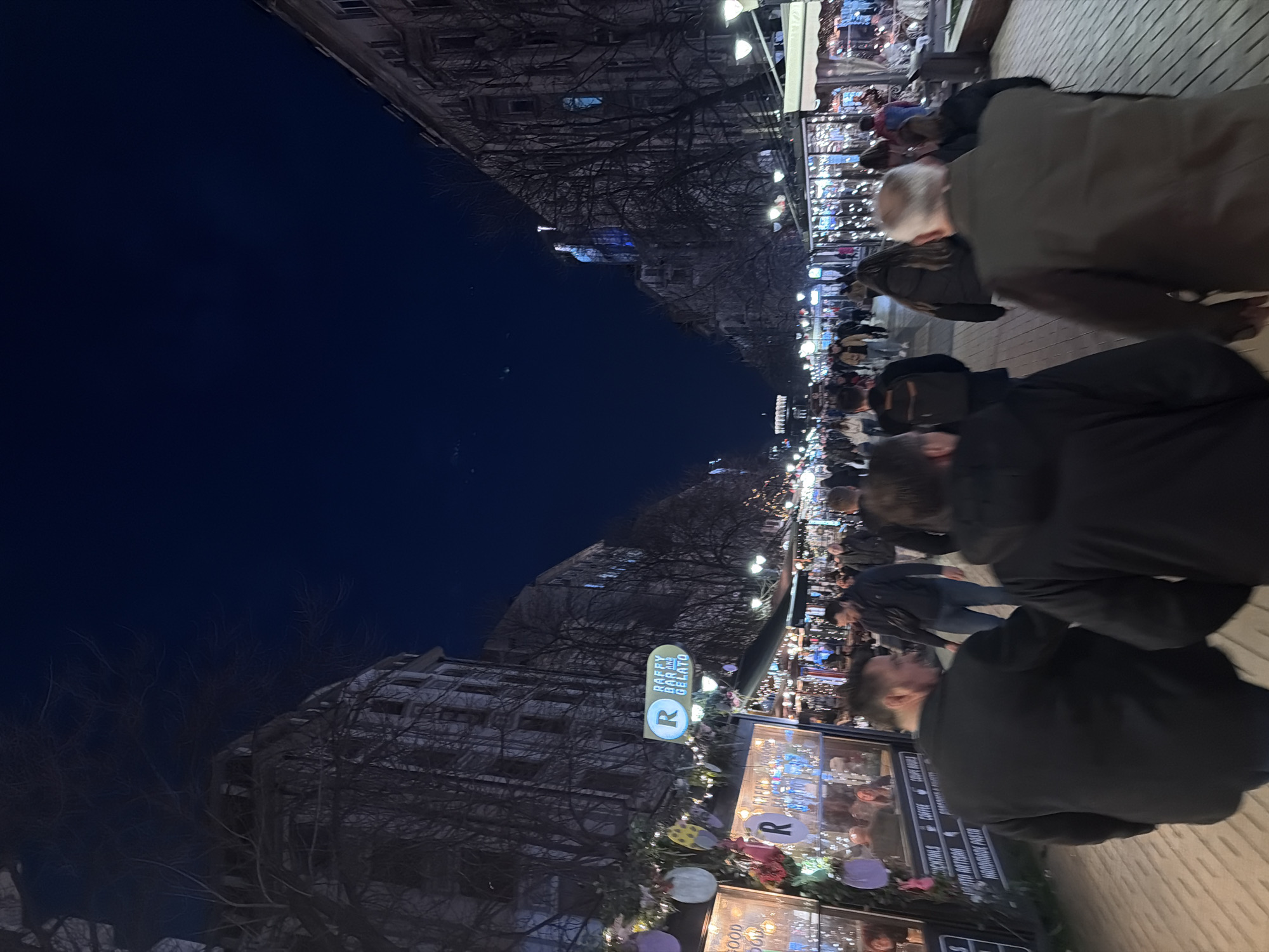 The image depicts a nighttime scene on Vitosha Boulevard, a pedestrian thoroughfare in Sofia, Bulgaria. The dark blue sky is visible above a long, straight street flanked by multi-story buildings exhibiting classical European architecture. Bare deciduous trees are interspersed along the street and against the building facades, indicating a cooler season.
The boulevard is artificially illuminated by numerous street lamps and lights from commercial establishments. Temporary market stalls or kiosks, adorned with string lights and festive decorations such as egg-shaped ornaments, line both sides of the street. A prominent sign on the left displays "RAFFY BAR AND GELATO" with a white 'R' on a blue disc and blue text on a yellow oval. Other visible signage on stalls includes terms like "COCKTAILS," "COFFEE," "COUPE BOWLS," and "HANDMADE PASTA." In the far distance on the right, a large, vertically oriented blue and white illuminated figure or projection is discernible on a building facade.
A substantial crowd of individuals, primarily adults, occupies the entire width of the paved walkway. The perspective is from behind several people in the immediate foreground, facing down the boulevard. Subjects are dressed in winter attire, including jackets and coats, and are observed moving in various directions along the crowded street. No specific interactions are clearly identifiable.