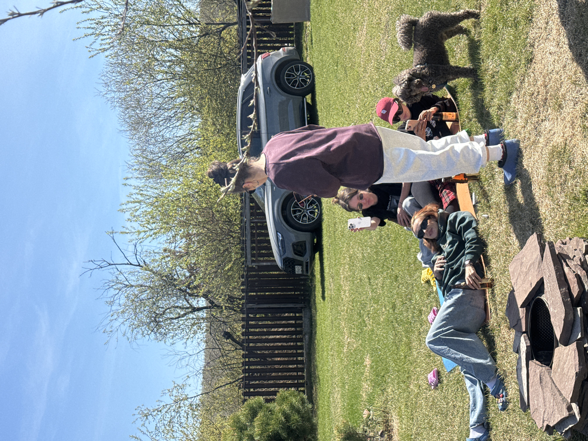 A group of people is sitting in a yard in Emel'yanovo, Russia. A gray SUV is parked near a wooden fence, and a brown poodle is standing on the grass. A woman in a maroon shirt and white pants is standing near a group of seated people. Another woman in a green hoodie and jeans is lying on the grass near a stone fire pit.