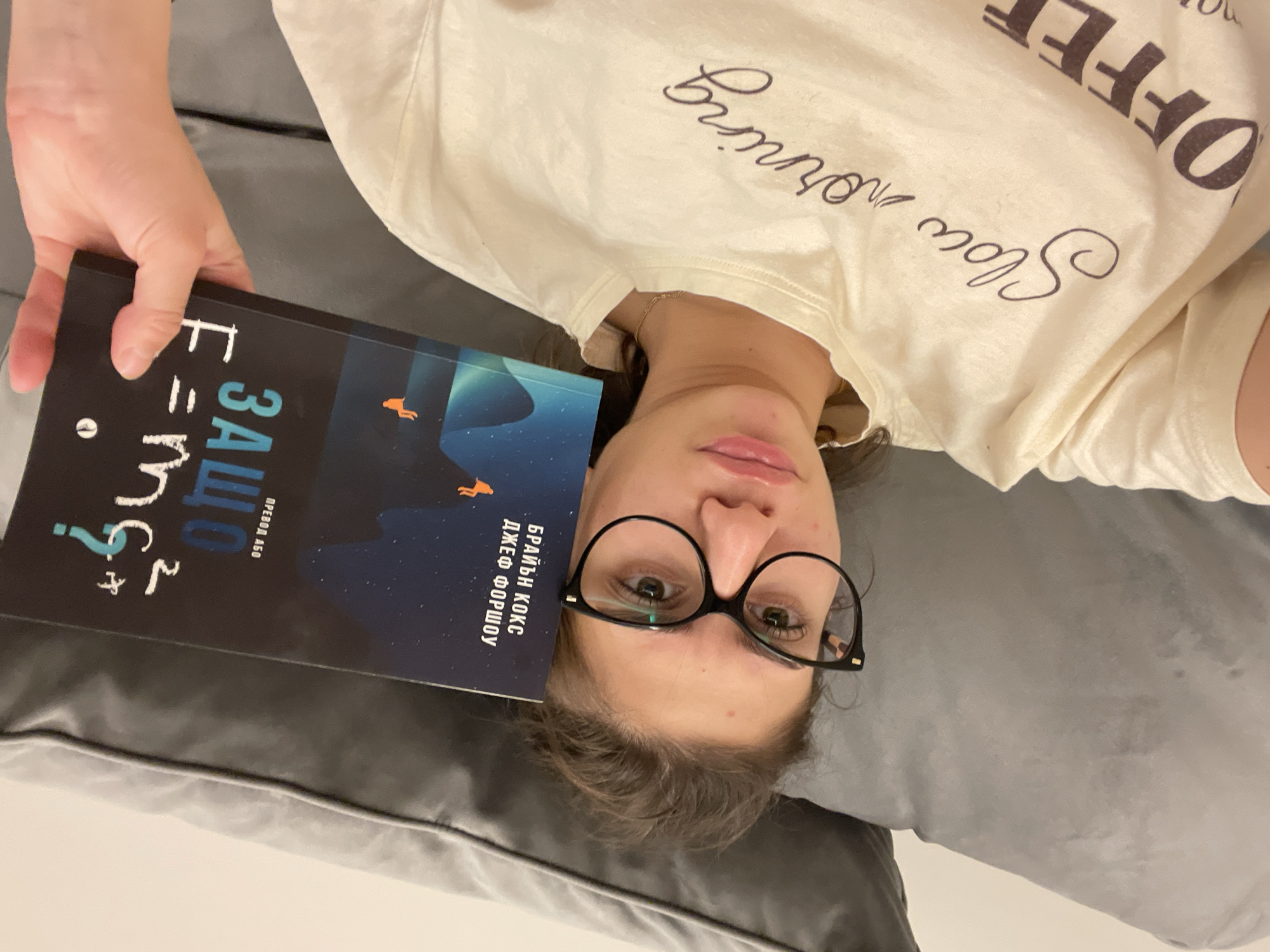 The image shows a woman lying on a gray pillow, holding a book titled "E=mc2". The book's cover is blue with an illustration of a starry night and an orange figure. The woman wears glasses and a cream-colored t-shirt with "Slow morning" and "COFFEE" printed on it. The image does not provide information about the location, nor does it mention Sofia, Bulgaria.