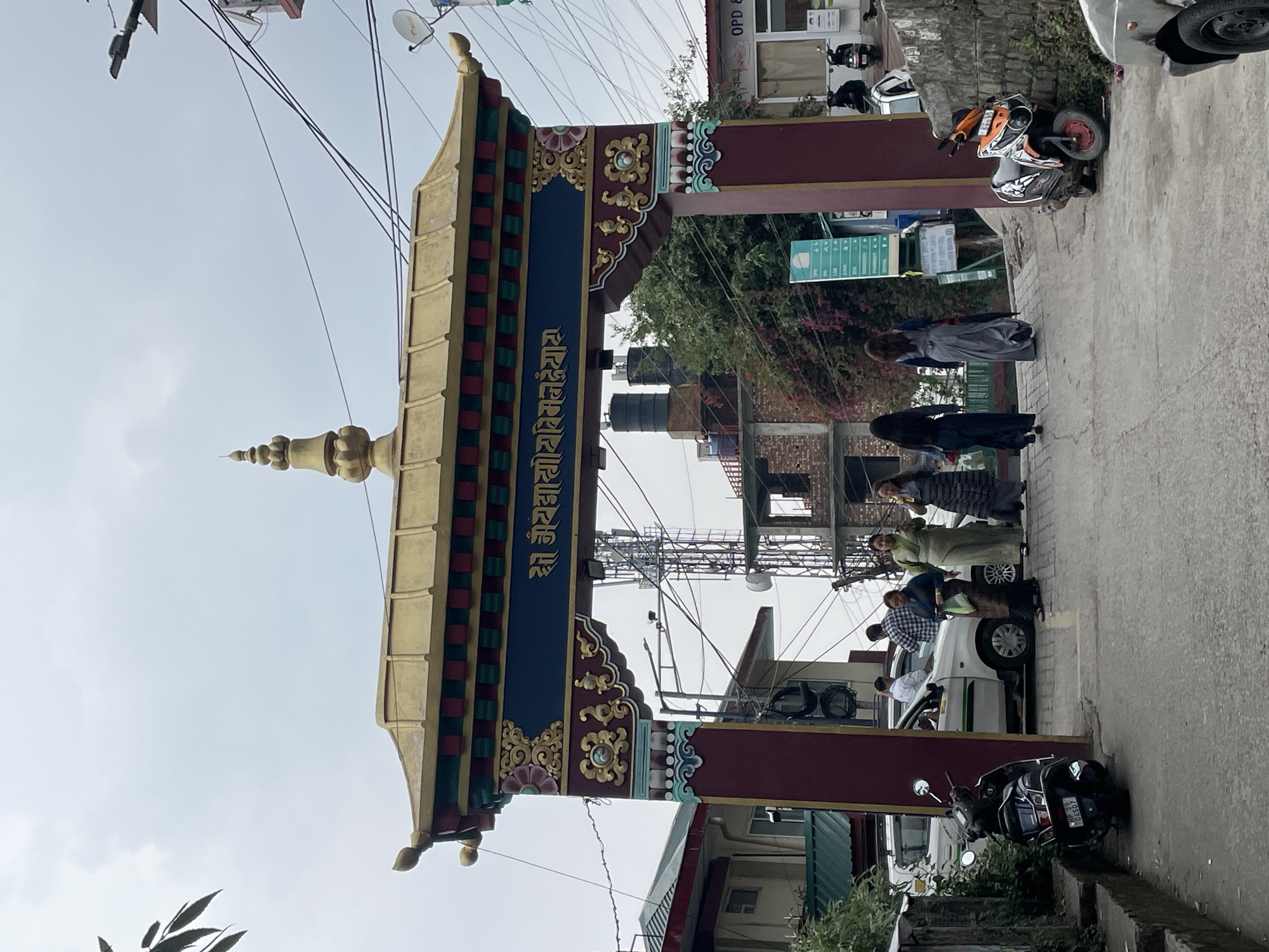A street view in Dharamshala, India, features a large, ornate archway constructed in the Tibetan style, embellished with a gold-colored roof, blue script, and red columns. The archway straddles a narrow street, with power lines overhead and various buildings visible in the background. Several pedestrians, including some wearing traditional Tibetan clothing, are walking along the street. Parked motorbikes and cars line the side of the road.