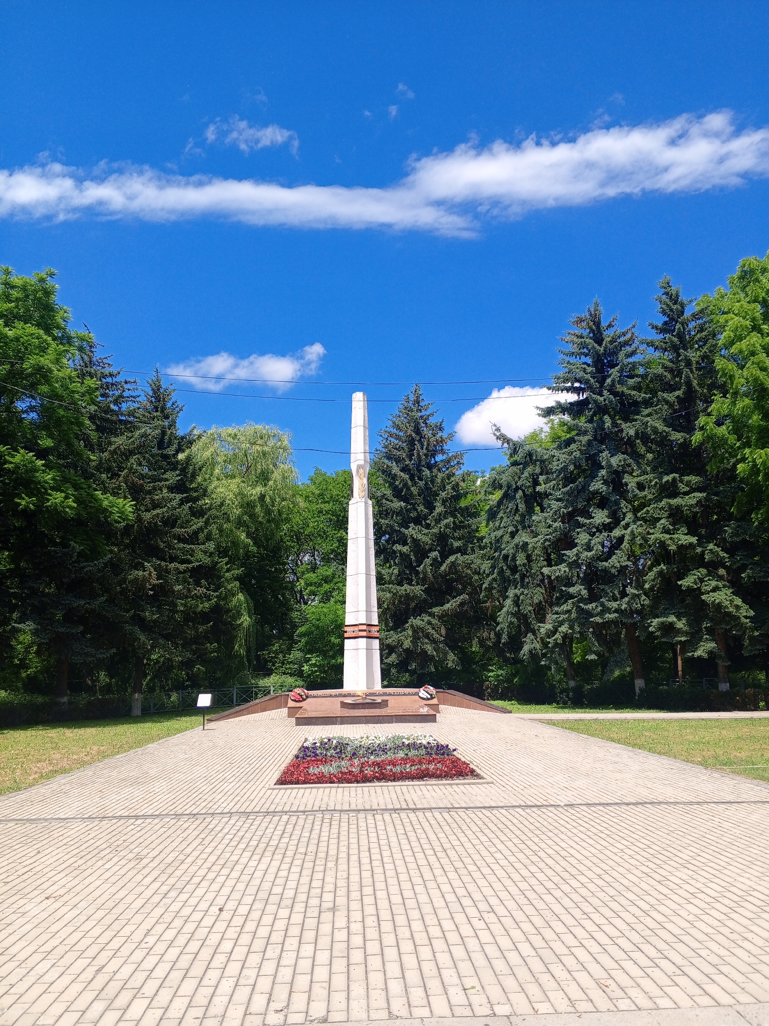 The image depicts a war memorial with a tall, obelisk-shaped monument in the center. The monument is white with some red and gold detailing, and appears to have an inscription or ornamentation at its base, though it is not clearly legible. In front of the monument is a flower bed with red and white flowers, and a wreath. The area around the monument is paved with light-colored rectangular stones, forming a symmetrical pattern. Flanking the paved area and the monument are grassy lawns and a dense line of evergreen trees, predominantly fir trees, with some deciduous trees interspersed. The sky above is a bright blue with scattered white, wispy clouds. The scene is set outdoors on a sunny day. Based on visual cues commonly associated with such memorials, and potentially correlating with other information if available, this could be a war memorial located in Yessentukskaya, Russia, likely commemorating fallen soldiers.