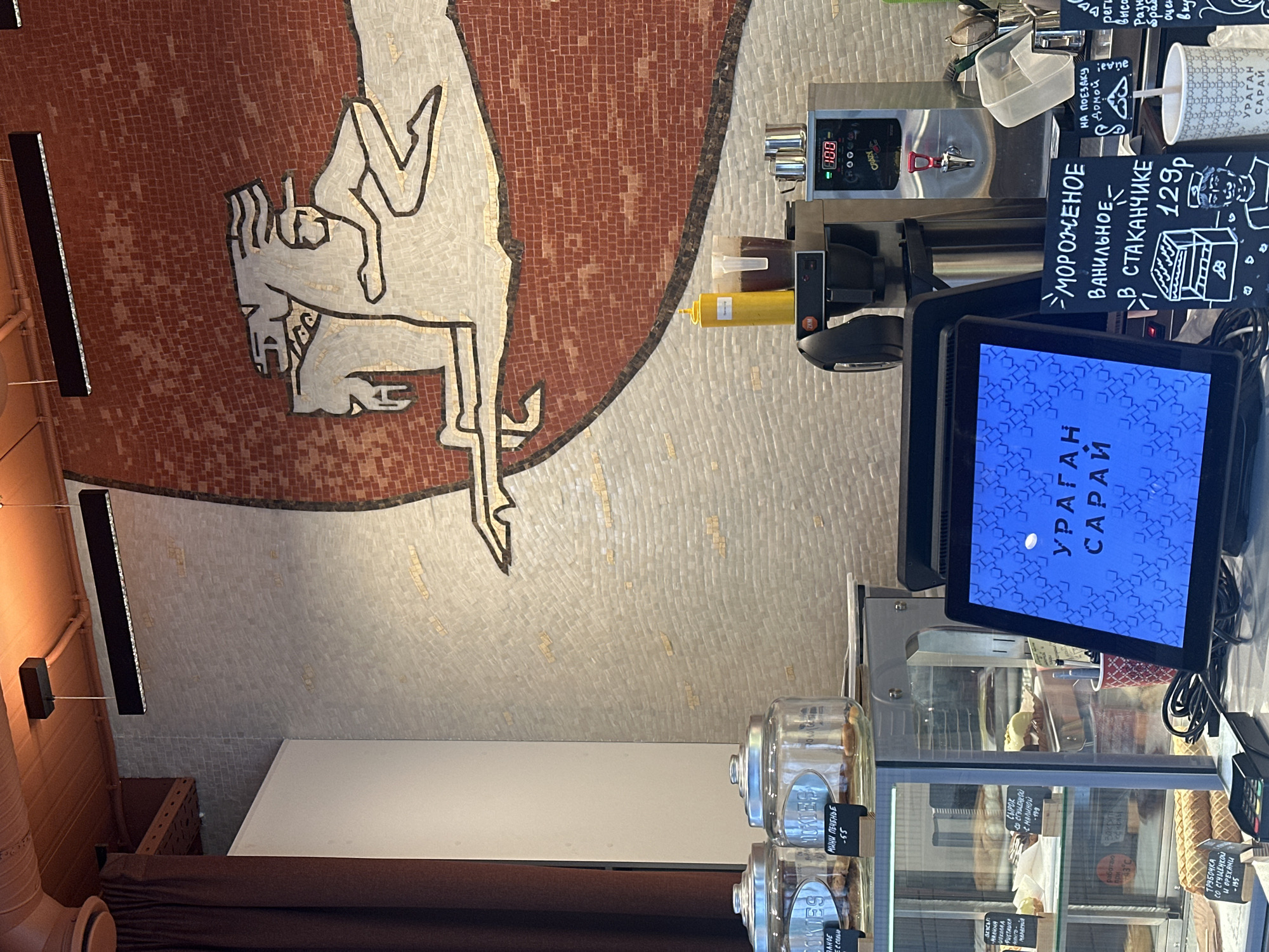 Here's a forensic description of the image:
**Location:** This scene appears to be inside a small cafe or shop, specifically in Kazan, Russia, based on the Russian language signage. The name of the establishment is likely "Ураган Сарай" ("Hurricane Saray").
**Environment:** The space is decorated with a mosaic mural on the wall, featuring a horse-like figure. The interior lighting seems warm, possibly from overhead fixtures.
**Objects:**
* A display case is filled with various baked goods and sweets, with price tags visible for items like mini cookies ("МИНИ ПЕЧЕНЬЕ -55") and pastries.
* Jars filled with cookies.
* A Point of Sale (POS) system with a screen showing the establishment's name.
* A drink dispenser, possibly for ice cream or coffee.
* Signage advertising vanilla ice cream ("МОРОЖЕНОЕ ВАНИЛЬНОЕ В СТАКАНЧИКЕ - 129p").
* Cups for take-away.
**Subjects and Actions:** The scene is static, with no people directly visible. However, the setting suggests that customers would typically be purchasing items from the display case and ordering drinks at the counter.