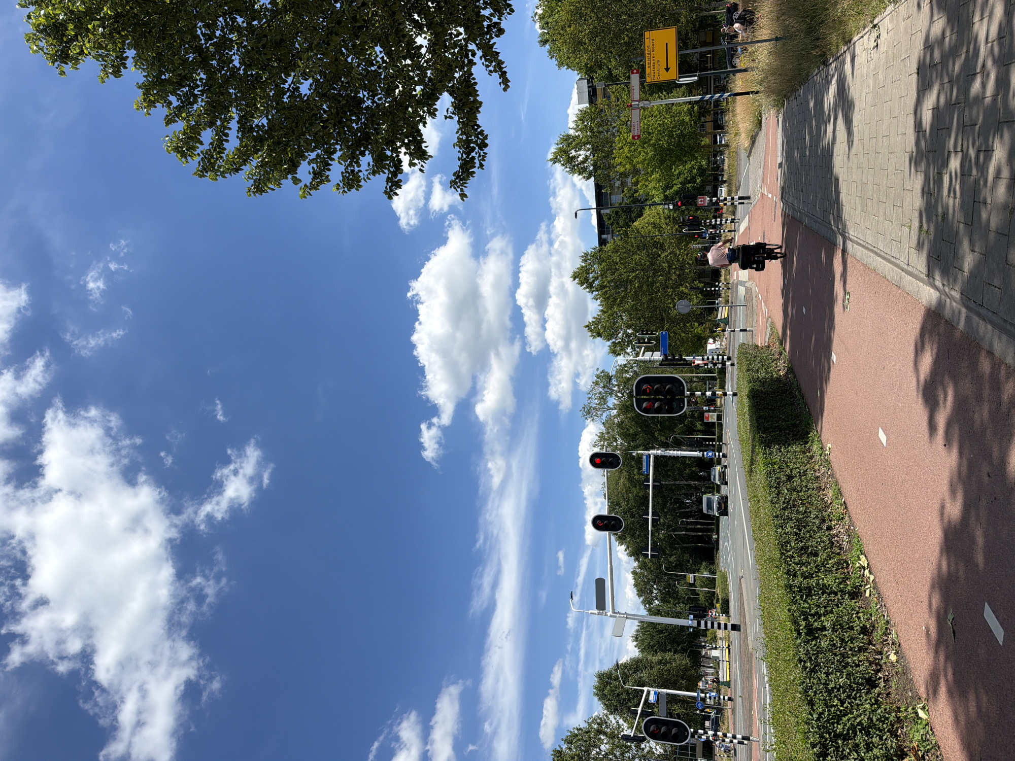 The image depicts a street scene in the city of Helmond, Netherlands, on a sunny day with a clear blue sky and scattered white clouds. A cyclist is riding on a dedicated red bike path, which runs parallel to a paved pedestrian walkway. The bike path is bordered by a manicured green hedge. In the background, traffic lights are visible, some showing red. Cars and a van are proceeding through an intersection. Trees line the street, and buildings are visible in the distance. A yellow directional sign with Dutch text indicating "omleiding" (detour) and "Houtsdonk-Noord Centrum" (Houtsdonk-North Center) with an arrow pointing left is mounted on a pole. A few individuals are seated near the walkway on the right side of the image.