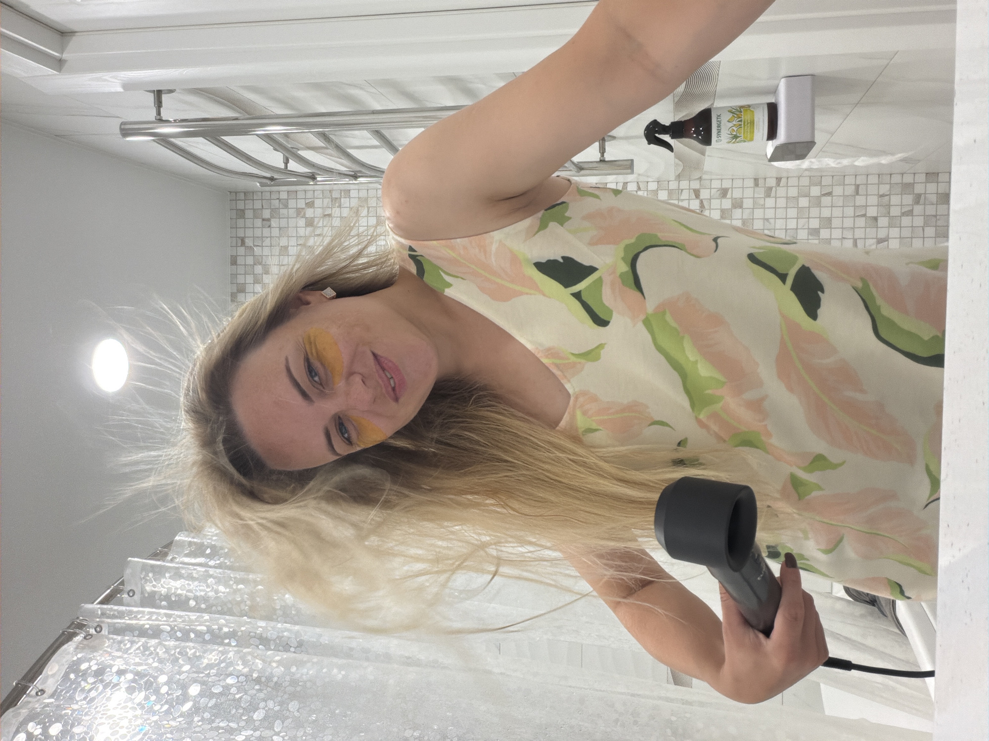 A woman with blonde hair, wearing yellow under-eye patches and a floral-print dress, holds a hair dryer in a bathroom. Her hair is being blown by the dryer, causing it to fly upwards. A shower curtain is visible behind her, and a towel rack is on the wall to her right. A bottle of cleaning spray sits on a shelf to the right of the towel rack. The location is not specified as Votkinsk, Russia.