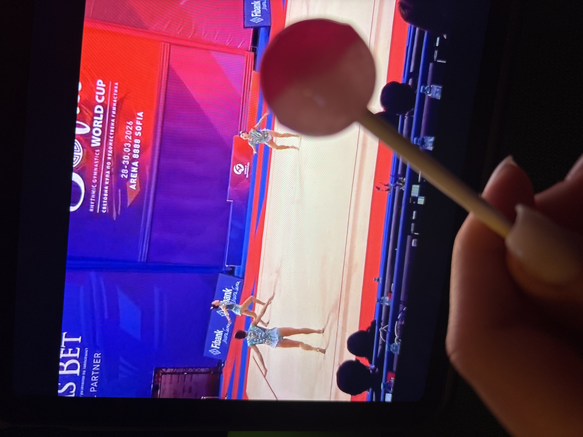 The image shows a screen displaying a rhythmic gymnastics World Cup event. The event is scheduled from March 28-30, 2026, and held in Sofia, Bulgaria, at Arena 8888 Sofia. Three gymnasts are performing on the floor. A red and white lollipop on a stick is partially obscuring the view. The tablet is being held in someone's hand.