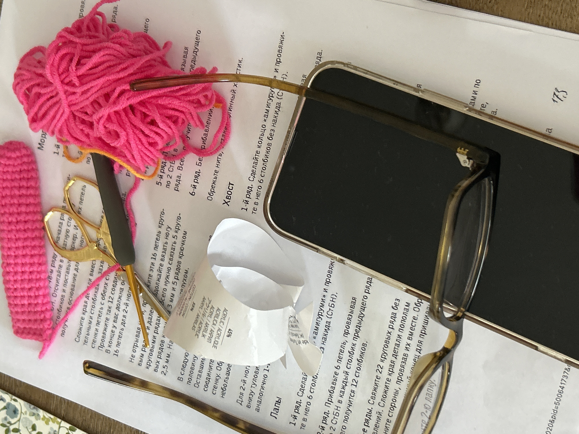 The image depicts a crafting project in progress, likely crochet or amigurumi. A small, pink crocheted object rests on a sheet of paper with handwritten instructions in Russian. Next to it is a ball of bright pink yarn and a pair of open scissors with black handles. A tortoiseshell-patterned crochet hook lies nearby. A pair of eyeglasses and a smartphone (screen-down) are placed over the instructions. A piece of torn paper is also on the instructions. The crafting materials are arranged on what appears to be a table.