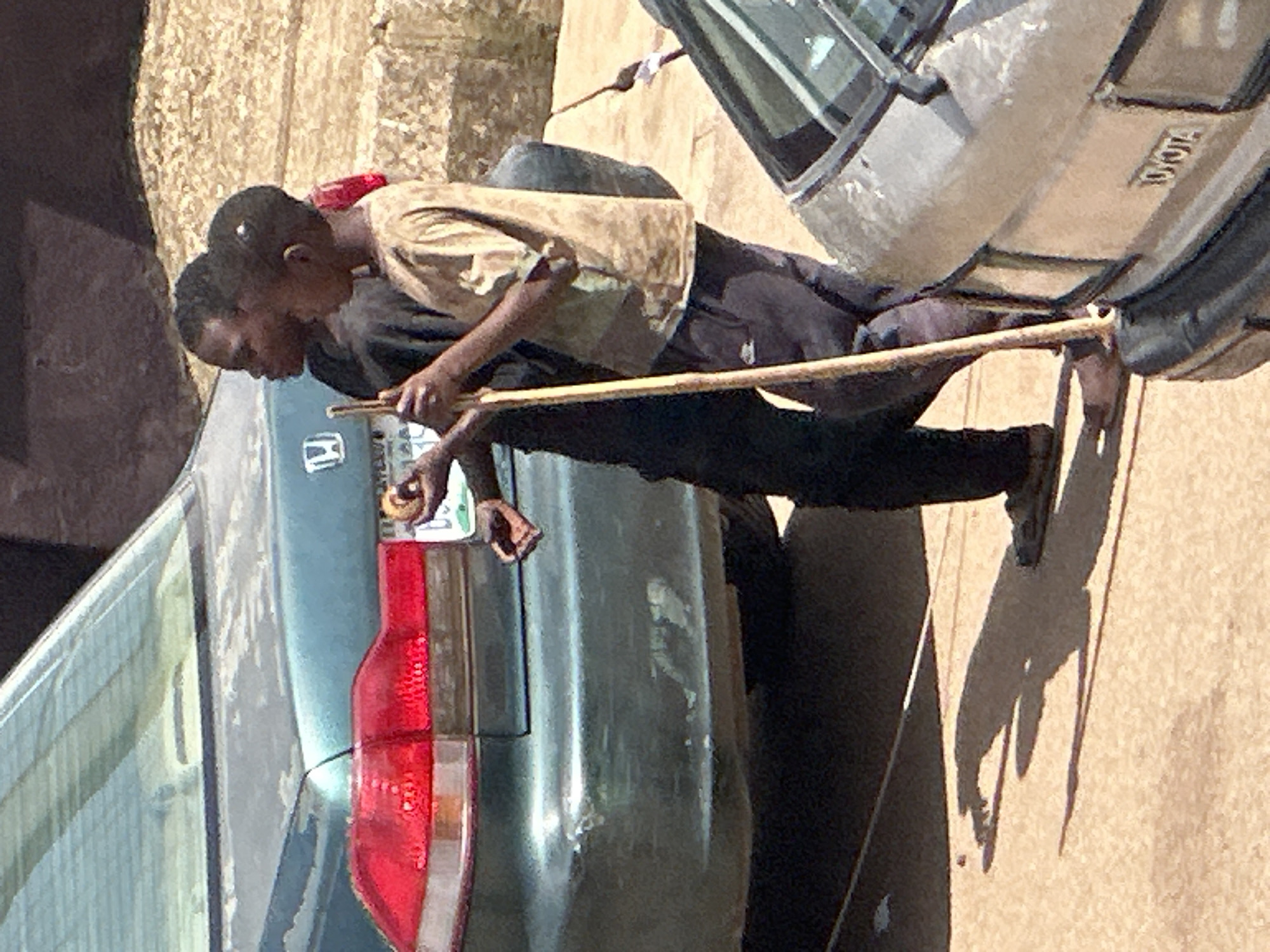 In Bukuru, Nigeria, two young individuals stand near vehicles. The foreground features the rear of a light-colored sedan, possibly a Toyota, with a visible "TOYOTA" emblem on its trunk. To the left, the rear of a dark teal sedan, appearing to be a Honda based on a visible emblem, is prominent. A young person in a tan shirt and dark pants holds a wooden stick upright, standing to the right of the dark teal car. Another young person in a dark shirt stands behind them, facing towards the dark teal car and holding a round, light-brown object. Both individuals are positioned on a paved surface, with shadows cast by the sun. The background is out of focus, showing a textured wall or structure.