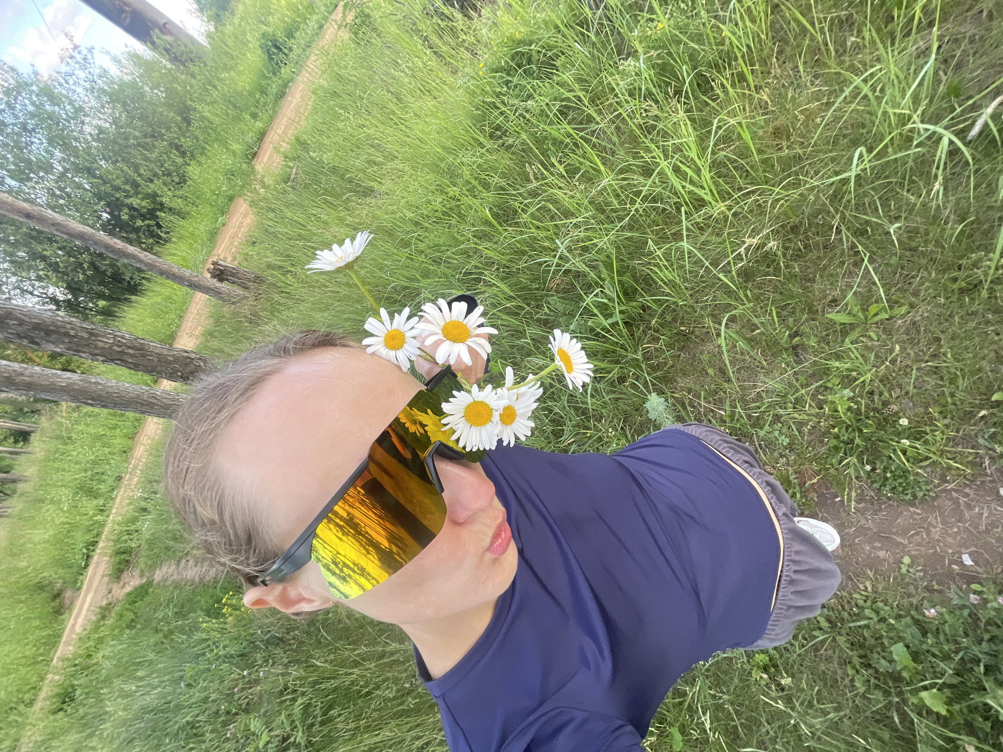 This is a high-angle, wide shot selfie taken outdoors in a grassy area, likely in Podshivalovo, Russia. The subject, a person wearing a dark blue t-shirt, grey pants with a yellow elastic band, and reflective sunglasses, is looking upwards towards the camera. Several daisies are placed around the subject's head, with some resting on their sunglasses. The background consists of lush green grass, a dirt path, and trees in the distance. The lighting suggests it is daytime, possibly late afternoon given the warm reflections in the sunglasses and the soft lighting. The overall mood appears playful and relaxed. There is no visible text in the image.