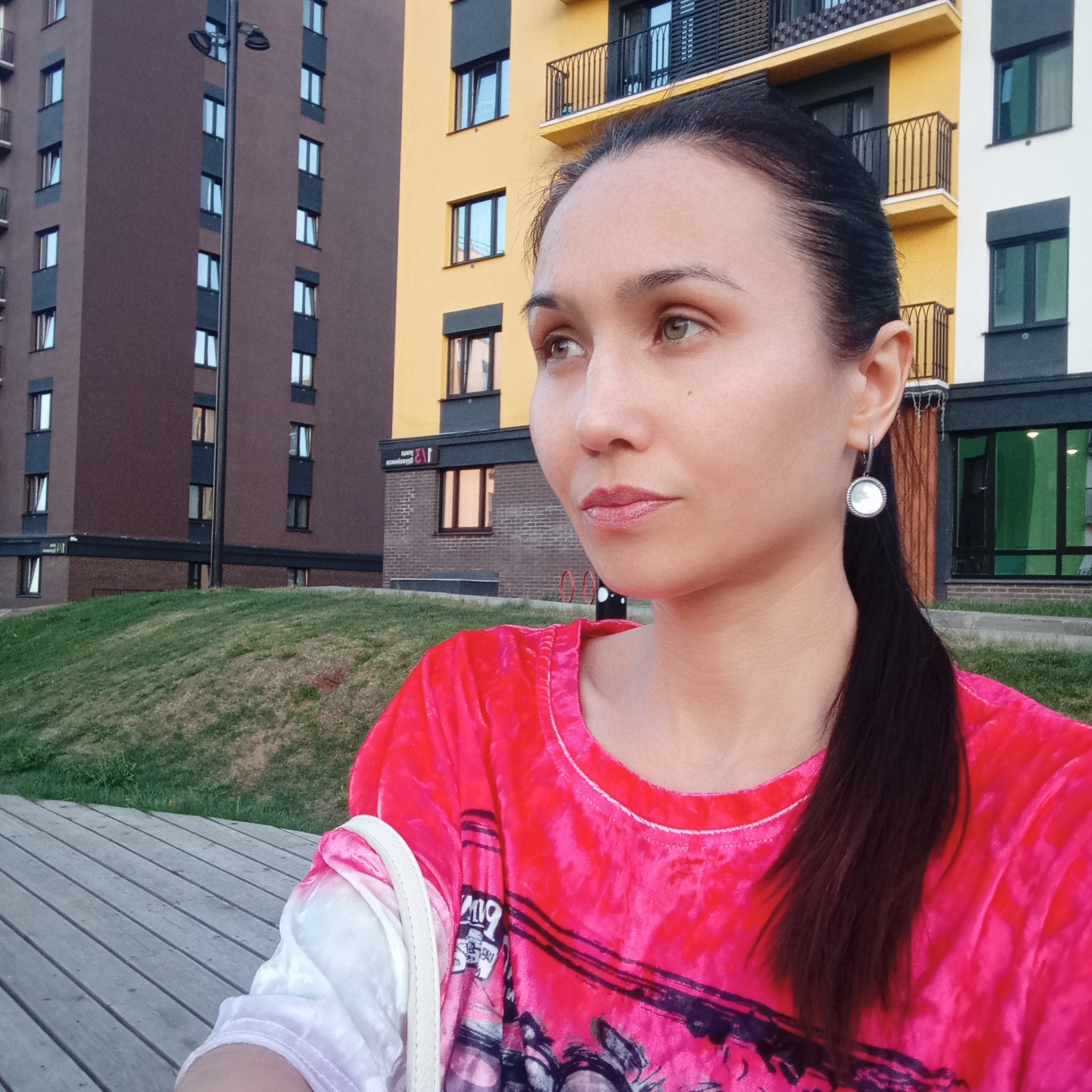 A woman with her dark hair pulled back into a low ponytail, wearing a vibrant pink t-shirt and a white shoulder bag, sits outdoors in Pervomaysky, Russia. She is looking off to the side with a contemplative expression. The background features modern apartment buildings in shades of brown, yellow, and white, along with a grassy area and a wooden boardwalk.