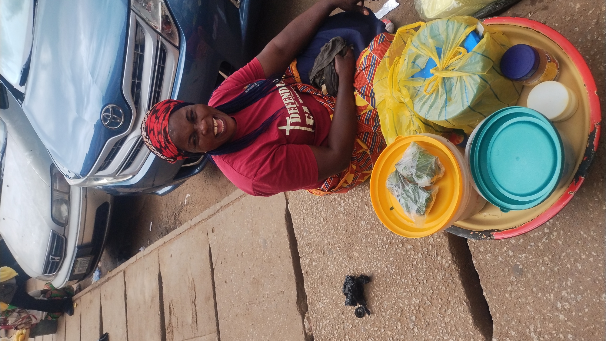 The image captures a smiling woman sitting on a paved surface next to a large tray of goods, with cars parked behind her. The woman, positioned prominently, wears a red short-sleeved t-shirt with partially visible white text that reads "DEFENDER" and features a cross symbol. She also wears a vibrant, patterned skirt in shades of yellow, red, black, and white, and a matching patterned headscarf. Her expression is one of broad happiness, as she looks towards the viewer.
To her right, a large, round, reddish-yellow metal tray rests on the ground. It holds several items, suggesting she might be a street vendor. These items include a yellow plastic bag containing a blue container, various lidded containers in blue and white, and a yellow plastic bowl holding several small, square-shaped food items wrapped in clear plastic. A turquoise container lid is also on the tray. On the ground near the tray, there is a small, dark, amorphous object, possibly a discarded item or another small bag.
In the background, two cars are visible: a blue Toyota with its front grille and logo clearly seen, and a lighter-colored car (possibly white or silver) behind it. The setting appears to be outdoors on a sunny day, likely on a sidewalk or street edge. The ground is made of light-colored, textured paving stones, with visible cracks.