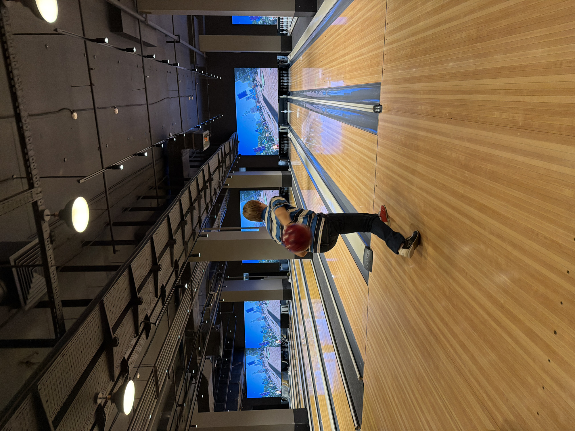 A young person is bowling in a modern bowling alley. They are mid-throw, with a red bowling ball in their right hand, blurred from motion, heading down a wooden lane. The person is wearing a blue and white striped t-shirt and dark jeans, with red sneakers. In the background, large screens display a virtual bowling scene with pins and animated characters. The bowling alley has multiple lanes, overhead lighting, and exposed ceiling infrastructure. The image appears to be taken from a slightly elevated angle, looking down the lane. The location of this scene is not specified in the image, and therefore cannot be definitively placed in Izhevsk, Russia.