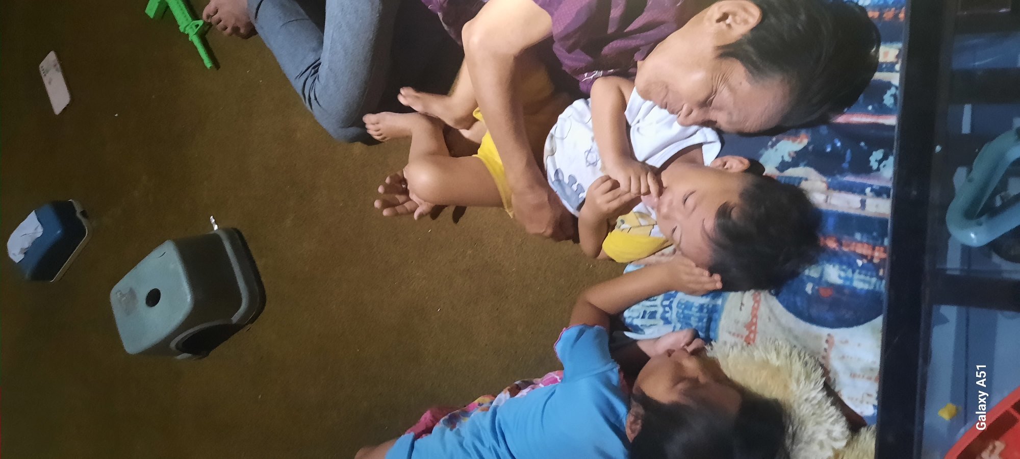 Three people, two children and an adult, are lying down on a brown carpeted floor. The two children are lying face to face, with their heads touching and their mouths open. One child is wearing a blue shirt and the other is wearing a yellow shirt and a white t-shirt. The adult is lying beside the children, wearing a purple shirt. In the background, there is a blue tissue box, a small green stool, a white rectangular object, and a green toy gun. The scene appears to be indoors. There is no indication of the city of Ploso, Indonesia, in the image itself.