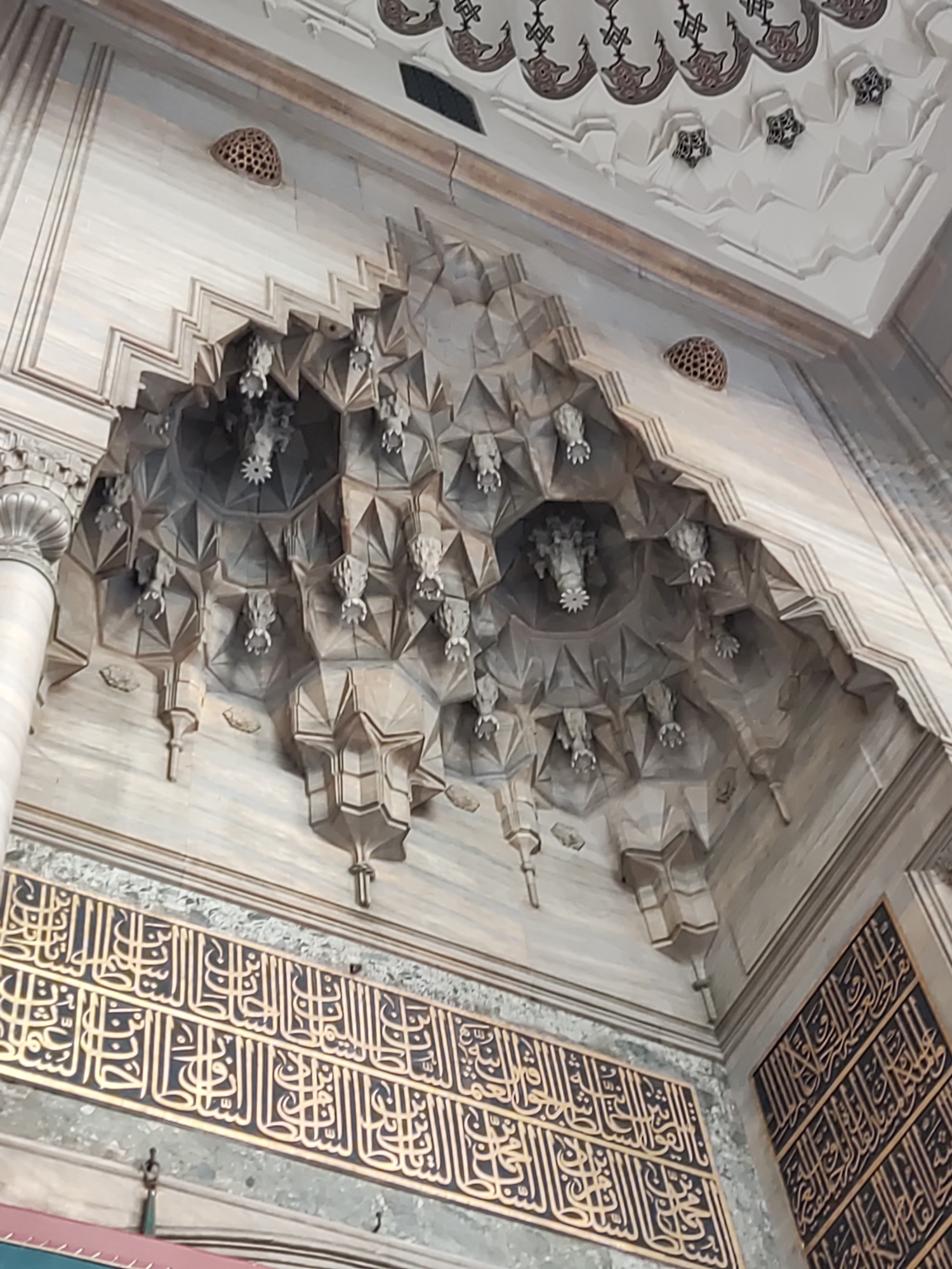 The image shows an interior view of a mosque in Istanbul, Turkey, likely the Sultan Ahmed Mosque (Blue Mosque) or the Süleymaniye Mosque, known for their intricate Islamic architectural details. The focus is on the elaborately decorated muqarnas (stalactite vaulting) ceiling and upper walls, rendered in carved stone with a light, marbled appearance. The muqarnas are arranged in a complex, honeycomb-like pattern with numerous small, inverted pyramid shapes, creating a three-dimensional, geometric effect. Small, decorative elements, possibly lamps or hanging ornaments, are visible within the recesses of the muqarnas.
Below the muqarnas, there is a band of ornate Arabic calligraphy inscribed on a dark background with gold lettering. This calligraphy likely consists of religious verses or decorative patterns. The calligraphy is arranged in horizontal rows. The walls themselves appear to be clad in polished marble, with subtle veining and variations in tone, contributing to the overall sense of grandeur and refinement. Two circular vents with patterned surfaces are visible in the upper wall area. The lighting appears to be a mixture of natural and artificial, highlighting the textures and details of the architecture. No people are visible in the image.