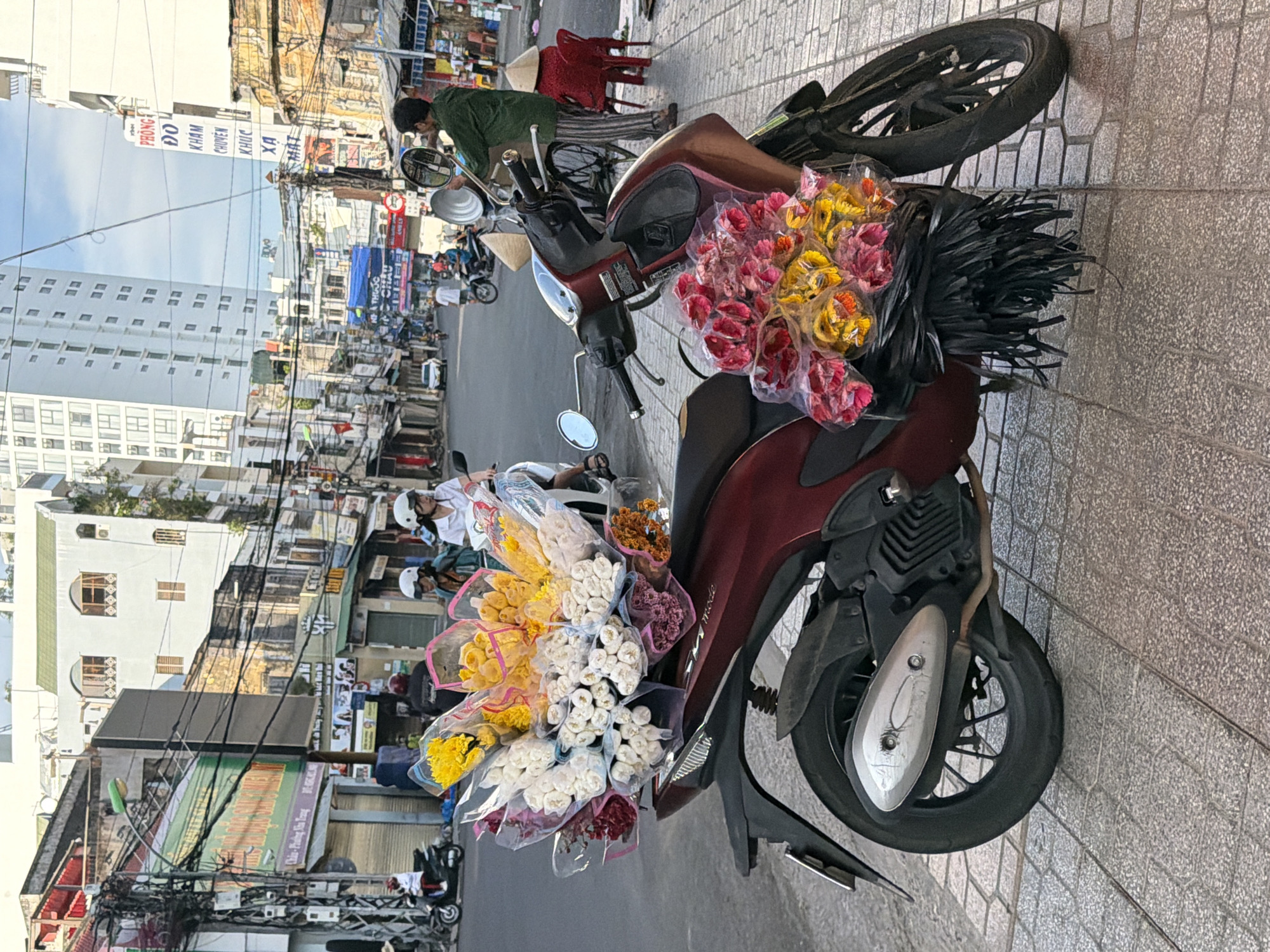 A maroon scooter is parked on a sidewalk in Nha Trang, Vietnam, loaded with large bouquets of flowers wrapped in plastic. The scooter's front basket contains bouquets of white, yellow, and pink flowers. The rear of the scooter also holds a bundle of pink and yellow flowers. In the background, a busy street is visible with other scooters, pedestrians, and buildings. A man in a green shirt stands near the scooter, holding a large metal lid. Red plastic chairs are set up on the sidewalk to the right, and several conical hats are visible. The environment suggests a typical street scene in a Vietnamese city, likely a marketplace or a street where vendors sell goods.