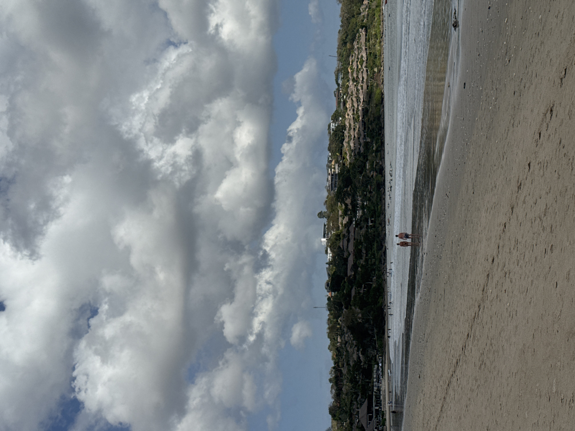 The image captures a tranquil scene at a beach in Kuta Selatan, Indonesia. In the foreground, a sandy beach stretches out, with the gentle waves of the ocean lapping at its edge. Two individuals, a man and a woman, are walking along the shoreline, appearing as small figures against the vast expanse of sand and sea.
Further into the image, the ocean appears calm with a few people visible in the water, possibly swimming or engaging in water activities. Beyond the water, a lush green hillside rises, dotted with numerous buildings that seem to be part of a resort or residential complex. These structures are characterized by their distinctive thatched roofs, blending harmoniously with the natural landscape. The sky above is dramatic, filled with large, white, and grey clouds, suggesting a partly cloudy day with potential for sunshine and shade. The overall atmosphere is serene and picturesque, highlighting the natural beauty of the Indonesian coastline.