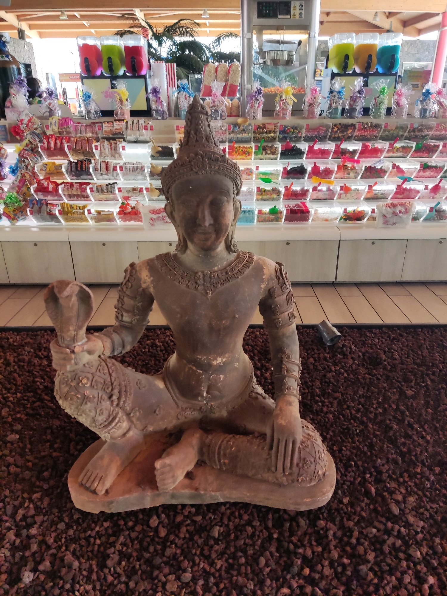 A weathered, tan-colored statue of a seated figure with a cobra entwined around its arm is positioned in the foreground on a bed of dark brown gravel. The statue faces forward, its left arm raised, holding the cobra which rears its head. The figure wears an ornate headdress and appears to be in a meditative or regal pose. Behind the statue, a brightly lit candy store is visible, with rows of clear plastic containers filled with various candies. These containers are arranged on white shelves, with more candies in bags and boxes behind them. A large dispenser holding three different colored slushy drinks is mounted on the back wall. The store appears to be indoors, with a wooden ceiling structure overhead. The scene is located in Adeje, Spain.