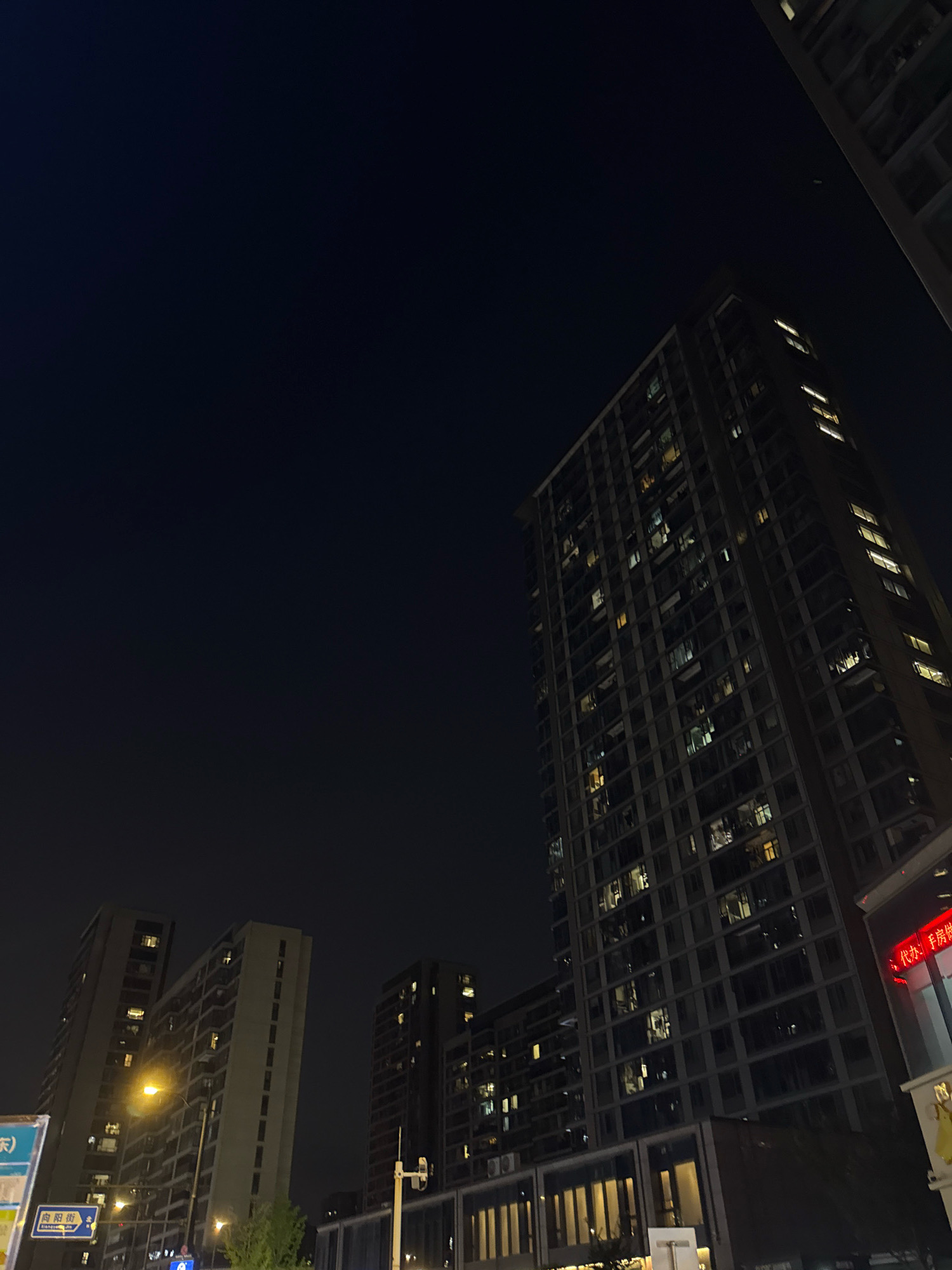 The image captures a nighttime street view in Yiwu, China, dominated by tall residential buildings illuminated by numerous lit windows. The buildings are modern, featuring dark facades and a grid-like pattern of windows. A street lamp with a warm glow is visible on the left side of the frame, casting light on the foreground. Signs with Chinese characters are present, one on the left with blue and white text, and another on the right with red text, suggesting commercial activity. The sky is dark, indicating the late hour. The overall impression is an urban environment at night, with the buildings and lights creating a sense of a bustling city even in the darkness.