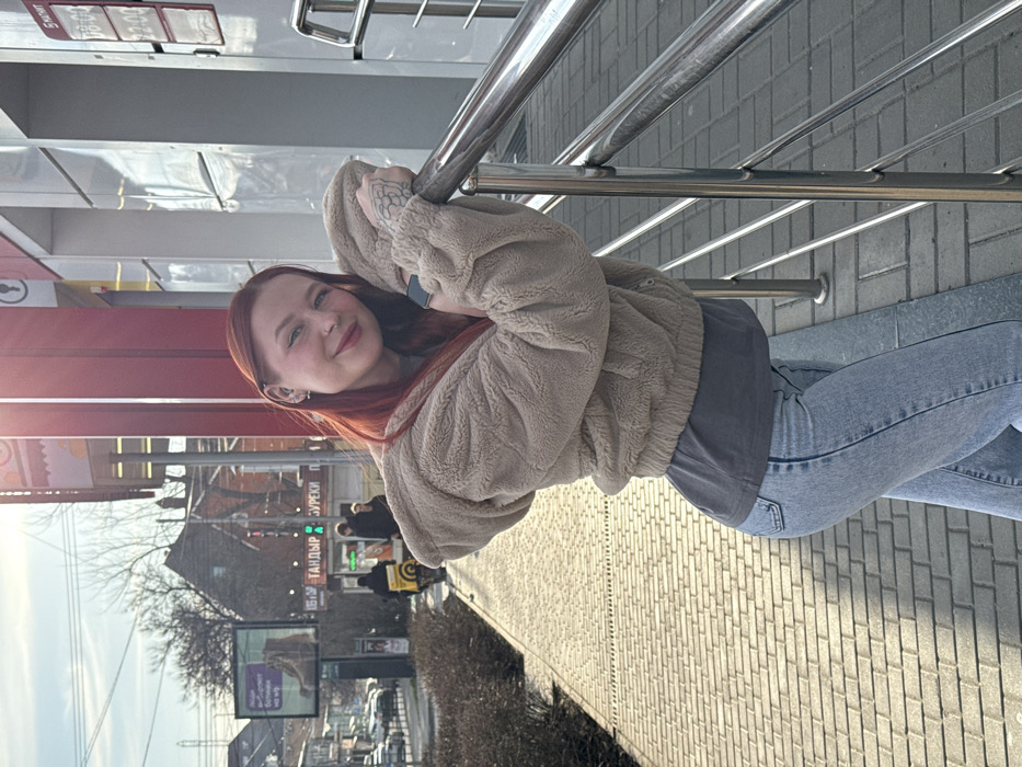 The image depicts an urban street scene in Rostov-on-Don, Russia, on a sunny day.
In the foreground, a young female with reddish-orange hair is positioned slightly left of center, facing the viewer with a subtle smile. She wears a beige hooded fleece jacket over a dark gray t-shirt and light blue denim jeans. Her left arm rests on a multi-barred chrome-plated railing, with her left hand gripping the top rail. A dark-colored smartwatch is visible on her left wrist, and a tattoo, resembling foliage, is present on her left forearm. She has multiple piercings, including a nose piercing and earrings in her left ear.
Immediately behind her is the entrance area of a commercial building, characterized by modern architecture with reflective surfaces and prominent red vertical paneling. A partially visible sign to her right indicates "5 МАТИ" and displays operating hours "08:00" to "22:00," identifying the establishment as likely a "Pyaterochka" supermarket. The ground in the foreground is paved with light-colored rectangular pavers.
The background reveals a bustling city street. To the left, a sidewalk continues, bordered by bare shrubs. Beyond this, a street with vehicles, including parked cars and one individual on a scooter with a large yellow delivery bag, extends into the distance. Building architecture varies, with some older brick structures featuring pitched roofs visible amongst more modern constructions. Bare trees suggest a late autumn or winter season. Several commercial signs are present, including a large billboard advertising footwear and a green illuminated sign displaying "ТАНДЫР БУРЕКИ" (Tandyr and Bureki), indicating a food establishment. Other pedestrians are intermittently visible in the background. Bright sunlight originates from the left, casting distinct shadows and creating lens flare in the upper left portion of the image.