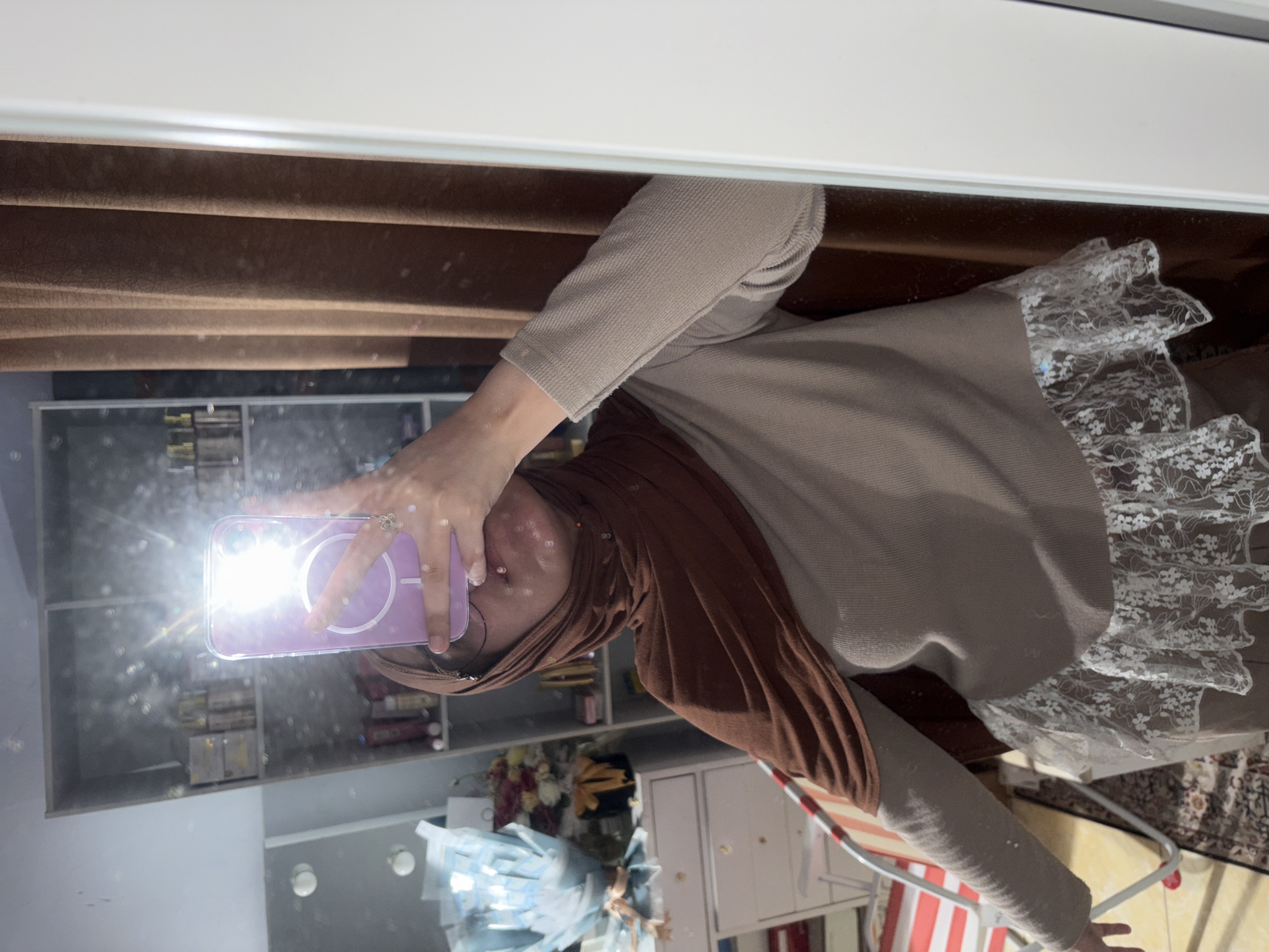 A person in a brown hijab and a beige long-sleeve shirt with lace trim is taking a mirror selfie indoors. The reflection shows shelves filled with various items, a bouquet of flowers, and a white cabinet. The person is holding a purple cell phone in their right hand. Background elements include brown window blinds and what appears to be a striped chair. There is no specific information to indicate the location being Gadingrejo, Indonesia.