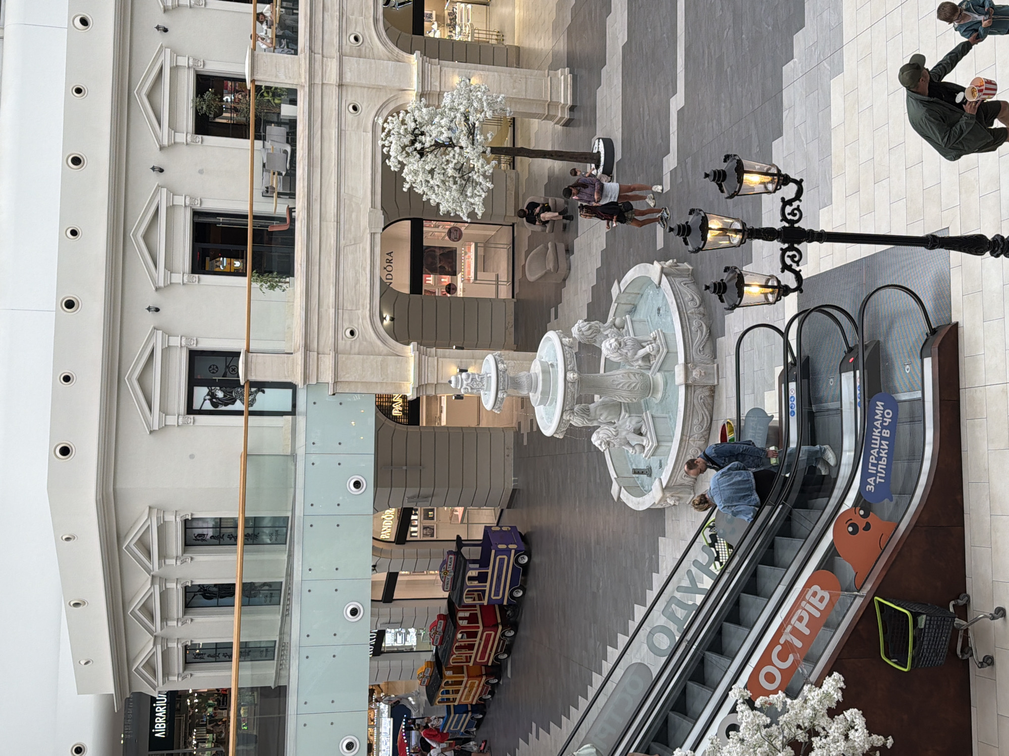 This is an eye-level shot of a shopping mall interior. The scene is bustling with activity, featuring escalators, shops, a decorative fountain, and people. The mall's architecture is grand, with ornate columns and a bright, airy atmosphere. In the foreground, an escalator is visible with people riding it, and a shopping cart is nearby. Further into the mall, a carousel-like train ride with colorful cars is set up, attracting children. A white artificial tree stands near a fountain with lion sculptures. Several shops are open, including a Pandora store. People are seen shopping, walking, and relaxing. The overall impression is one of a lively and modern commercial space. The text on the escalator signs indicates the location is likely in Kyiv, Ukraine, with the word "ОСТРІВ" (Ostriv, meaning "island") and other Ukrainian text visible.