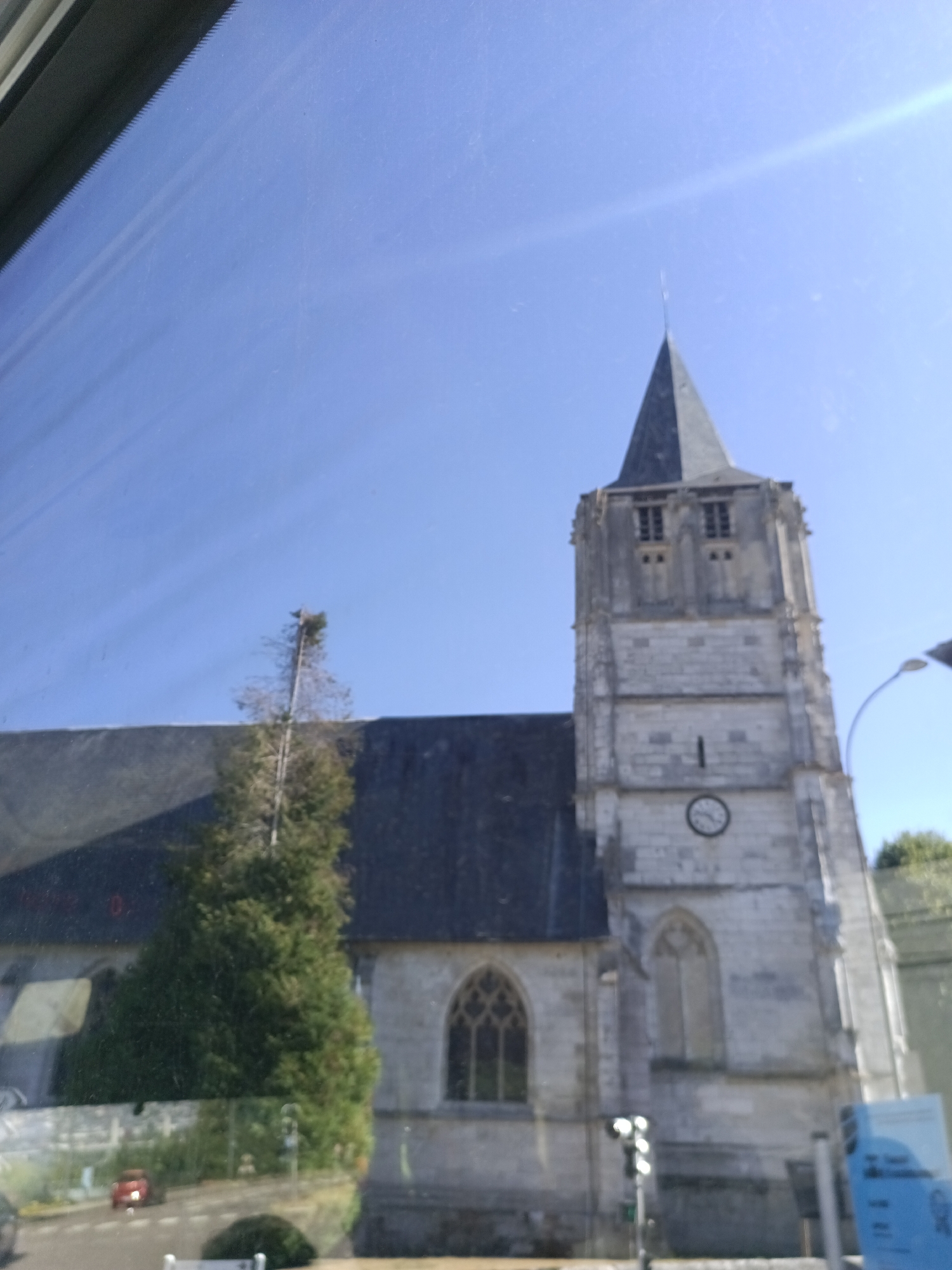 The image shows the exterior of a stone church with a tall spire in Canteleu, France. The church has a dark-tiled roof and Gothic-style windows. A clock is visible on the tower of the church. In the foreground, a tree is situated in front of the church. The scene is viewed through what appears to be a window, with reflections visible on the glass. The sky is clear and blue. A street light and some greenery are also present in the background. A red car is visible in the distance, driving on a road.