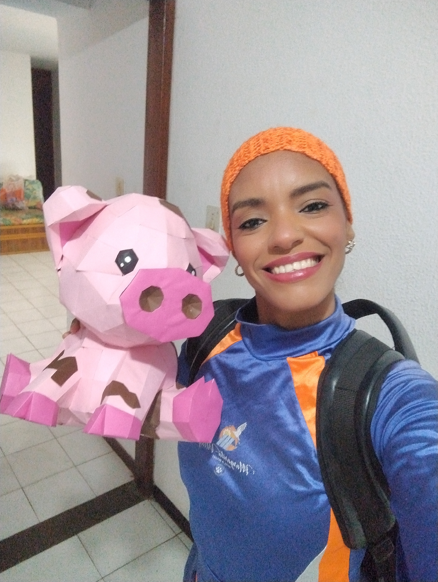 A woman poses for a selfie indoors, holding a pink, polygonal paper pig. She wears an orange knitted cap, a blue and orange shirt, and a black backpack. She is smiling. The background shows a tiled floor and a partially visible room with furniture. The location is likely an indoor space in Porlamar, Venezuela.
