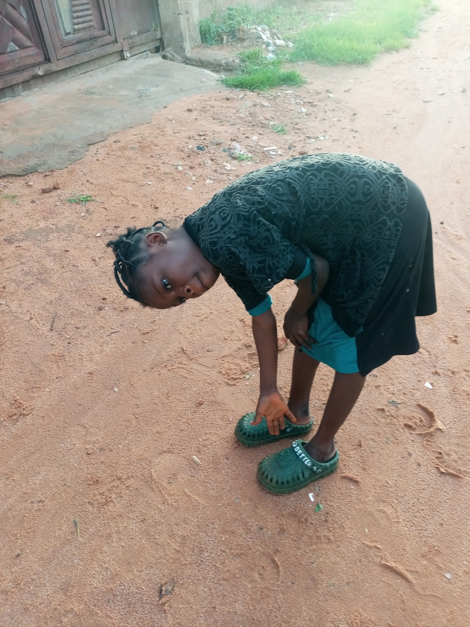 A young child, appearing to be of Nigerian descent, is pictured in a dusty outdoor environment, likely in Tempure, Nigeria. The child, wearing a dark, patterned top over teal shorts and green clog-style sandals, is bent at the waist, looking towards the camera. The ground is covered in reddish-brown sand, with scattered debris and patches of dry grass. In the background, a concrete structure with wooden doors is visible, suggesting a residential or commercial area. The lighting indicates it is daytime.