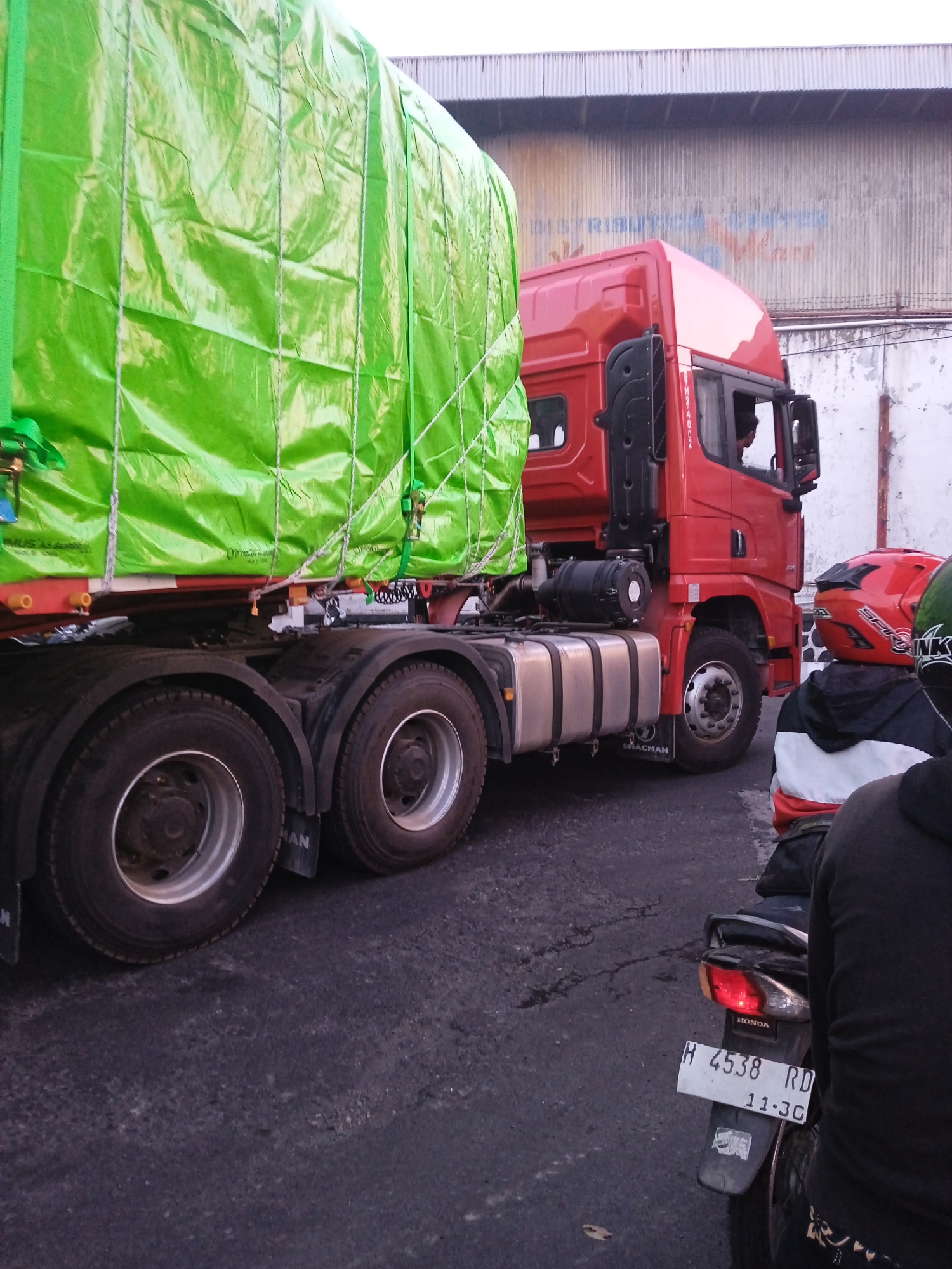 A red semi-truck with a bright green tarp-covered cargo is parked on an asphalt road, possibly in Semarang, Indonesia. The truck has dual rear wheels and the name "SHACHAN" is visible on the tire fender. In the foreground, a motorcycle with a visible license plate reading "H 4538 RD 11-3C" is parked. Two people are on the motorcycle, one wearing a red helmet and the other a black jacket with white and red accents. A weathered building with a corrugated metal roof and partial text on its facade is visible in the background. The sky is overcast.