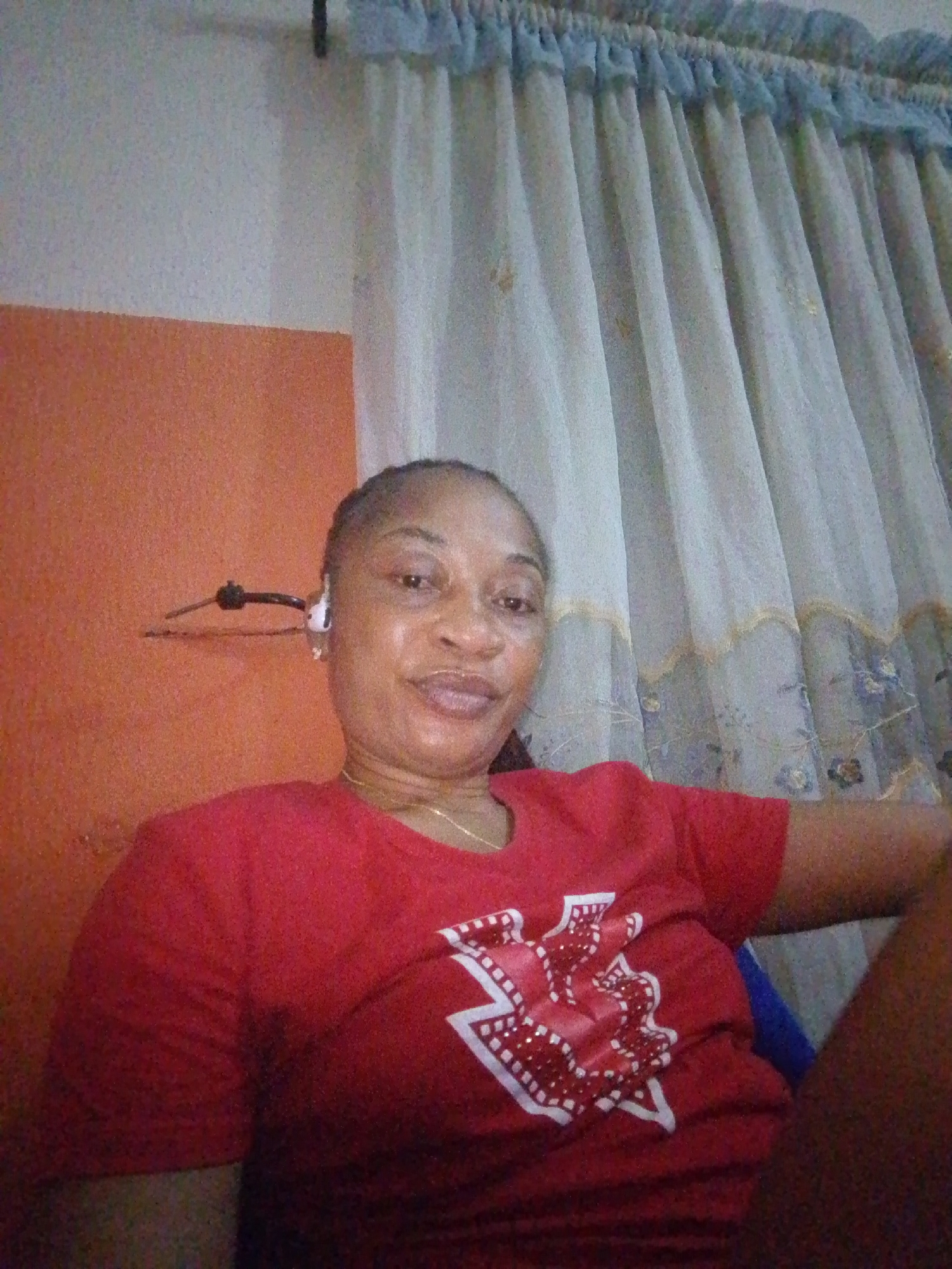 A medium shot, taken from a slightly low angle, shows a Black woman with braided hair and wearing a red t-shirt. She is looking at the camera with a slight smile and her mouth open. She is wearing a gold necklace. Behind her is an orange wall and a white curtain with a blue trim. A black wire is visible on the orange wall, and it appears to be connected to her earbud.
