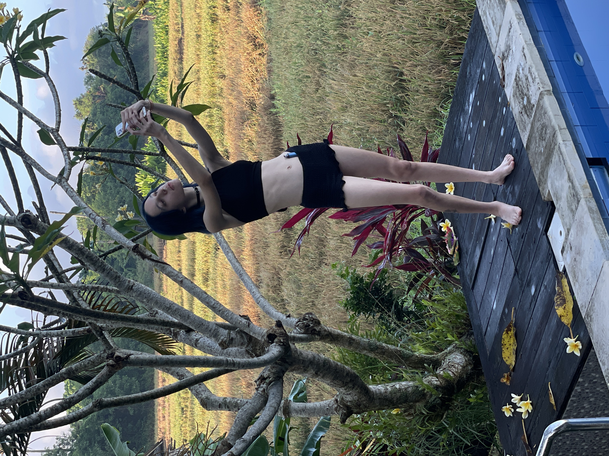 The image captures a young woman in Mengwi, Indonesia, lying on a dark wooden platform adjacent to a pool, evidenced by the blue tile edging. She is dressed in a black swimsuit and holds a smartphone above her, presumably taking a selfie. Her hair is dyed blue, and she is barefoot. The setting includes a verdant rice field in the background, a tree with bare branches in the foreground to the left, and tropical plants with red leaves near the platform. Fallen yellow flowers are scattered on the wooden surface.