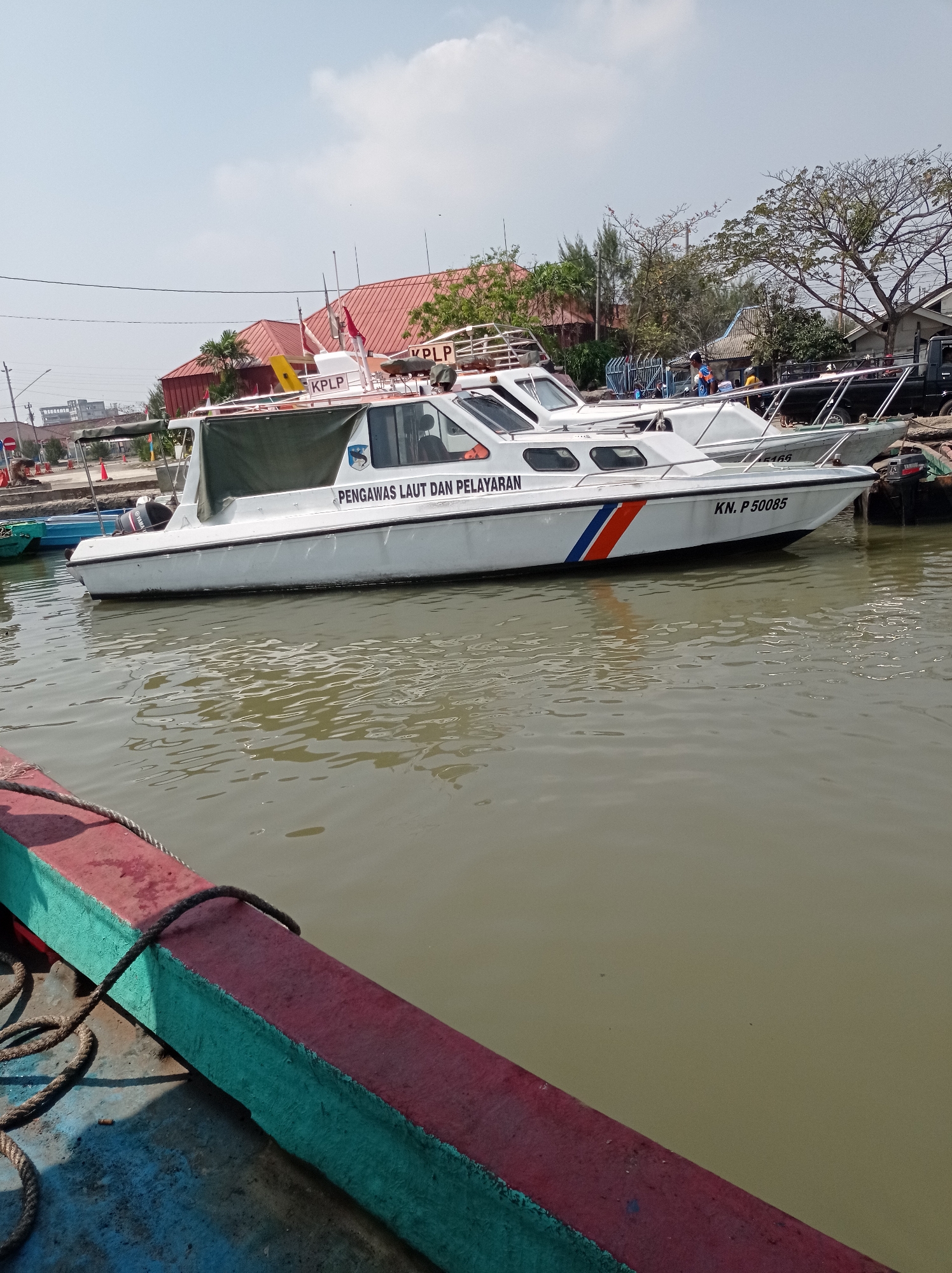 In Tegal, Indonesia, a bright, sunny day casts shadows as several boats are docked. In the foreground, a thick, dark rope is tied to the edge of a vessel, its weathered blue and green hull contrasting with a striking red band. Further in the water, a white patrol boat named "PENGGOWAS LAUT DAN PELAYARAN" with the identification number "KN. P 50085" is moored. The boat features a dark green canopy and the letters "KPLP" are visible on its roof. Another similar boat is partially visible behind it. The background reveals buildings with red roofs, trees, and a clear blue sky with sparse clouds.
