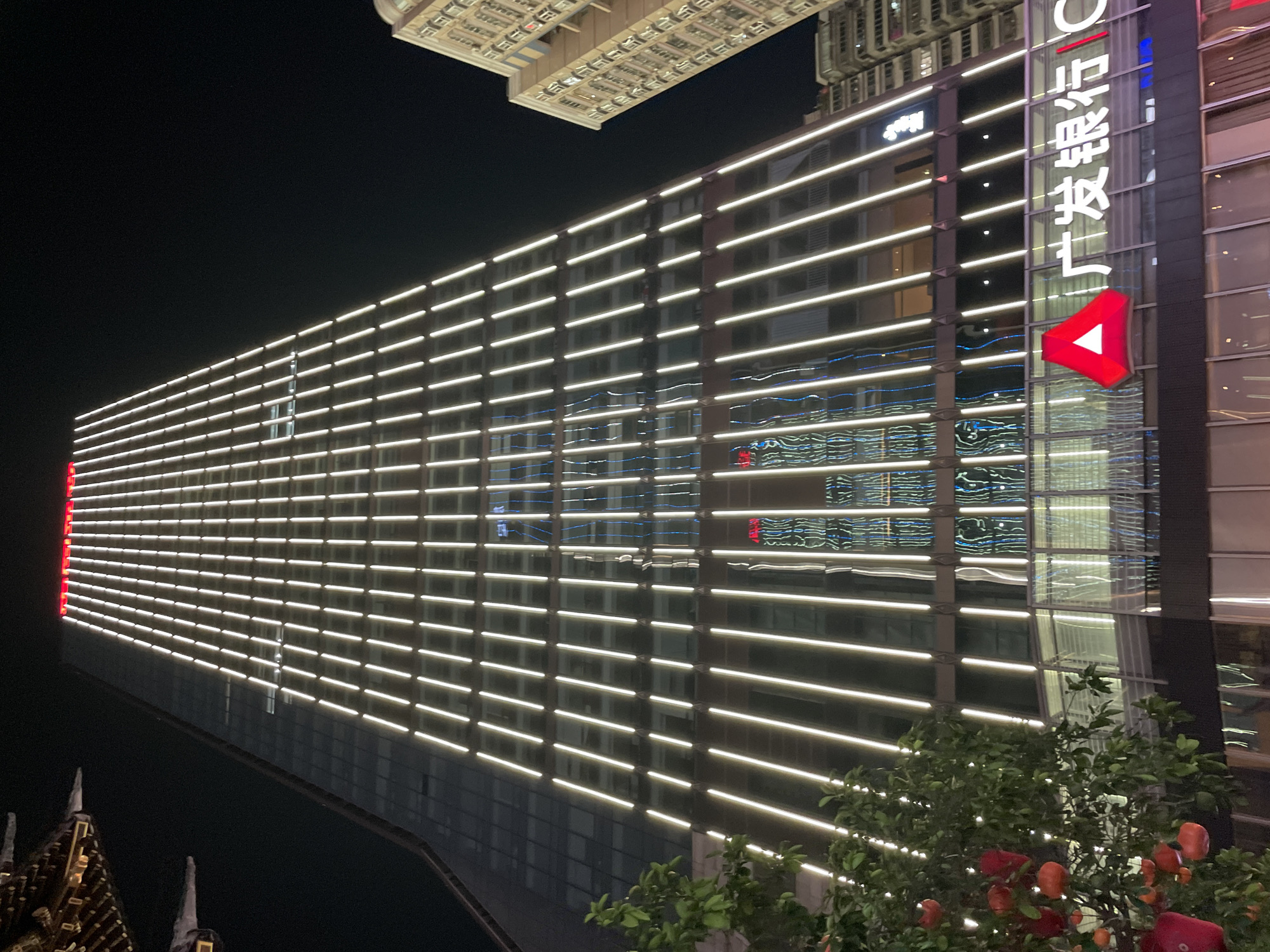 The image captures the nighttime exterior of a modern, illuminated skyscraper in Yuzhong District, China. The building is characterized by its sleek glass facade and vertical white LED lighting that accentuates its height and structure. At the top of the building, a band of red lights is visible. Reflections of colorful lights, appearing as blue and green patterns, are visible on the glass panels, suggesting the presence of other illuminated buildings or digital displays in the vicinity.
At the base of the skyscraper, a prominent logo featuring a red triangle within a white circle, accompanied by Chinese characters that spell out "Guangfa Bank," is displayed. This indicates the building likely houses the Guangfa Bank. To the right of this main skyscraper, another multi-story building with a more traditional architectural style and numerous illuminated windows can be seen. In the foreground to the left, a tree with green leaves is adorned with several bright red ornaments, possibly decorative lanterns, which add a splash of color to the scene. The overall environment appears to be an urban setting at night, with the architecture and lighting dominating the visual.