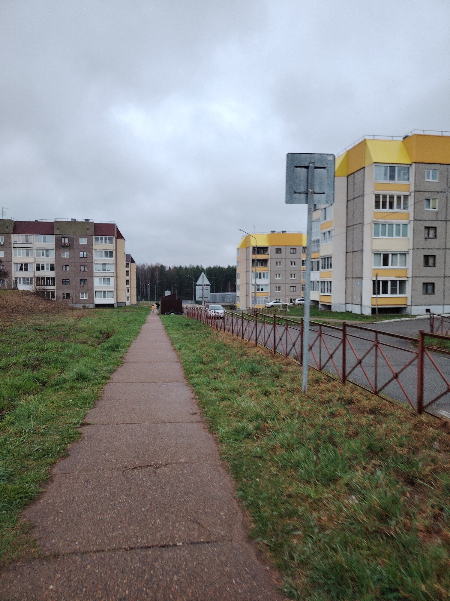The photograph depicts a street scene in Markovskii, Russia. A paved sidewalk stretches into the distance, flanked by grassy areas on both sides. On the left, there is a multi-story residential building with a red roof. On the right, a similar building has yellow accents. A brown metal fence runs along the right side of the sidewalk, and a grey sign stands next to it. Cars are parked in front of the building on the right. In the background, a line of trees can be seen. The sky is overcast and grey. There are two people walking in the distance.