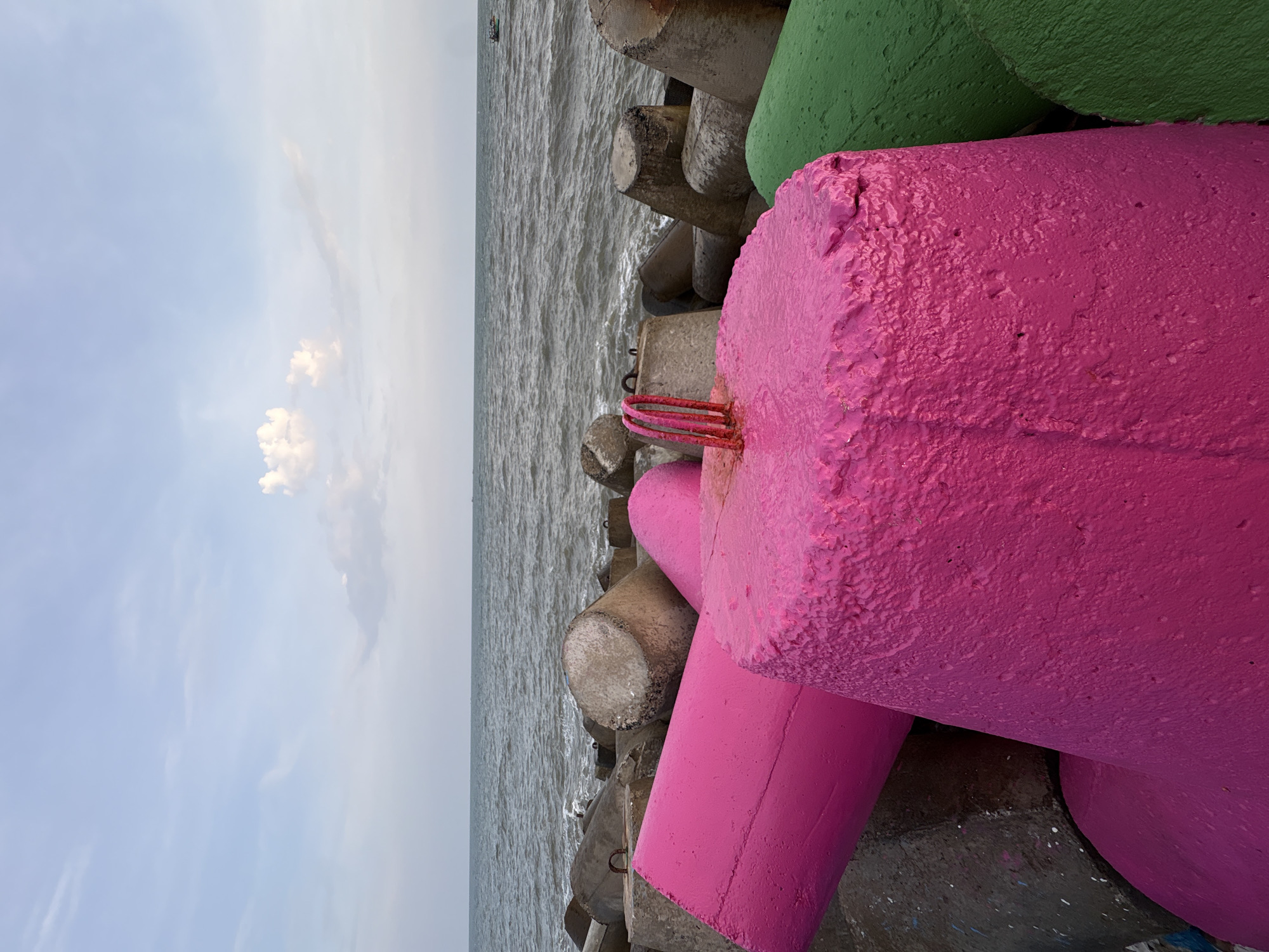 The image captures a section of a breakwater on a coastline in an unknown location in Vietnam. The foreground is dominated by several large, cylindrical concrete structures, some of which are painted in bright, contrasting colors. Two prominent pieces are painted a vibrant pink, with one in the immediate foreground having a rough, textured surface and a cluster of red wire loops protruding from its top. Another pink cylinder is angled diagonally behind it. To the right, a green cylindrical structure is partially visible.
Behind these brightly colored elements, a jumble of unpainted, weathered concrete tetrapods and cylinders form the bulk of the breakwater. These structures are rough and show signs of age and exposure to the elements.
The sea stretches out behind the breakwater, with moderate waves creating white foam near the shore. The water is a muted gray-blue color. In the distance, a small, dark boat is visible on the horizon.
The sky above is a pale blue with scattered white clouds. The lighting suggests it may be late afternoon or early evening, with a soft, diffused light. The overall scene is one of industrial coastal defense meeting artistic intervention, with the colorful concrete elements standing out against the natural backdrop of the sea and sky.